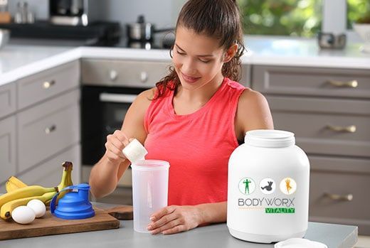 Woman mixing protein powder in a shaker cup in kitchen, bananas and eggs nearby.