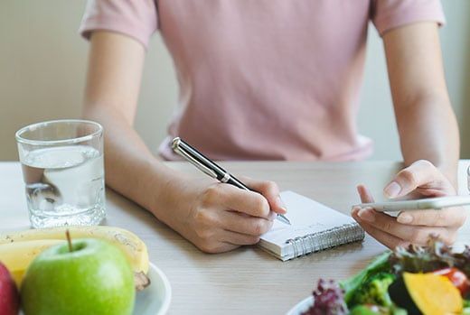Person writing in a notepad, with fruit, a glass of water, and a salad on a table.