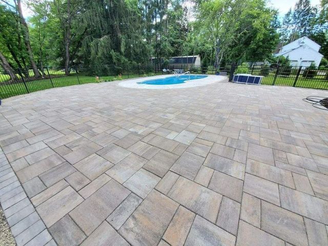 Patio pavers beside a backyard swimming pool, bordered by trees and a white shed.
