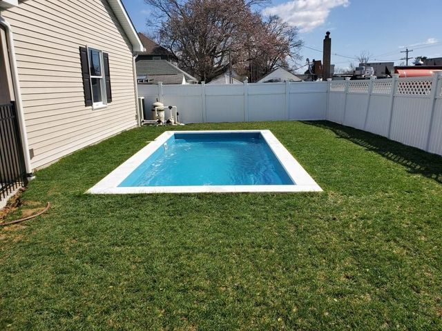 Backyard with a blue rectangular pool, green lawn, and white privacy fence beside a beige house