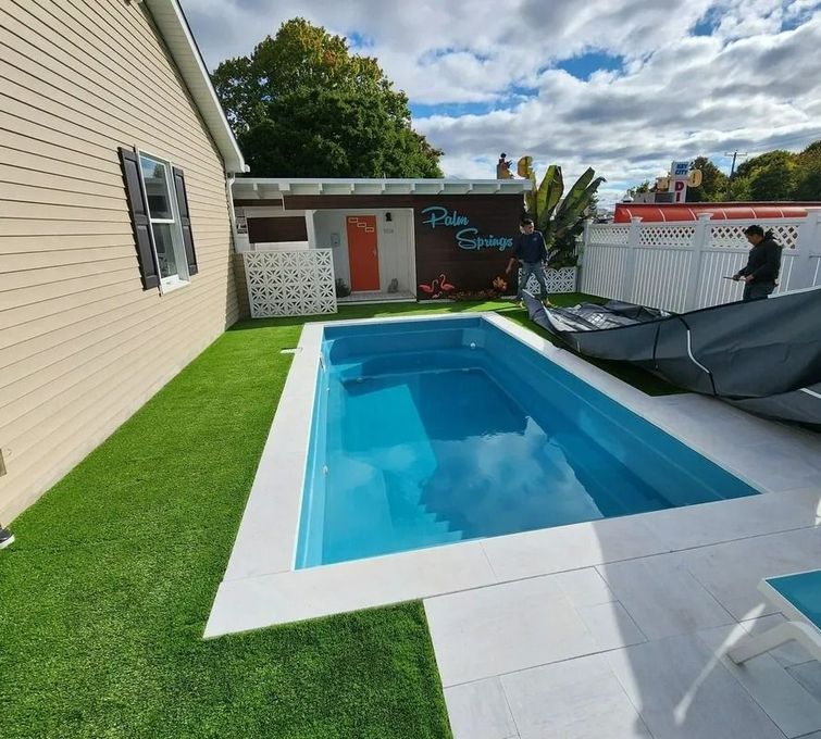 Backyard pool beside a beige house, with green turf, patio tiles, and a covered lounge area.
