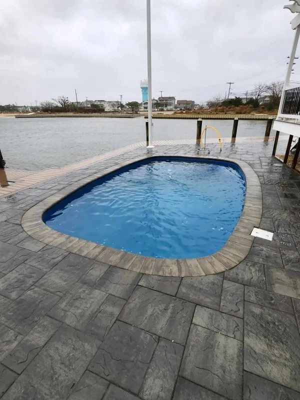 Small blue pool beside a rainy waterfront walkway with gray stone tiles and a cloudy sky