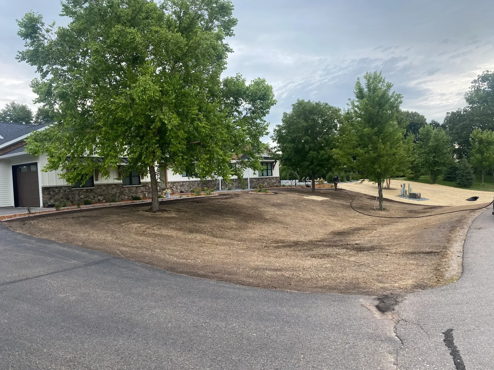 Curving driveway beside a house with trees and a dry dirt yard under a cloudy sky
