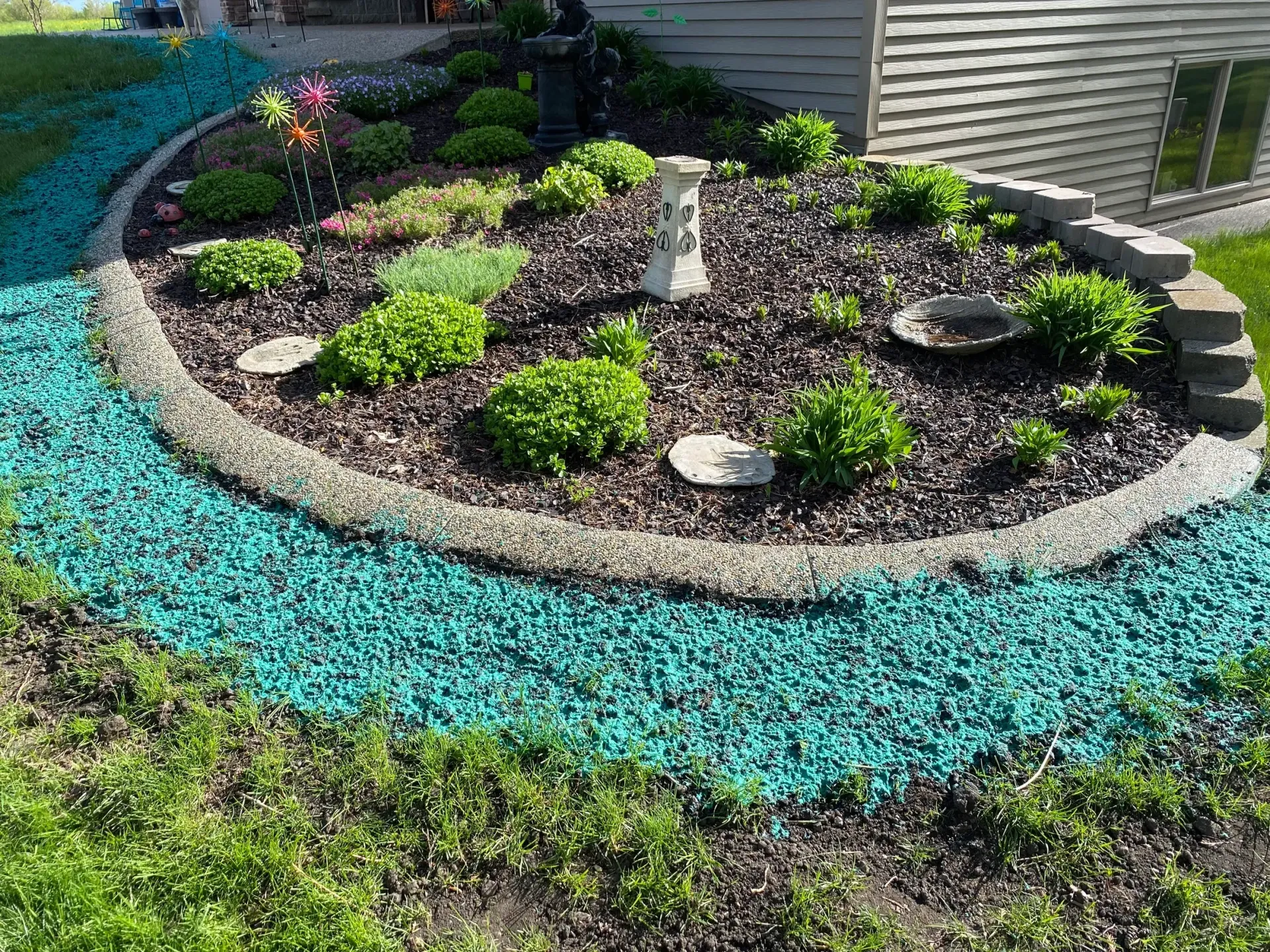 Circular flower bed with small shrubs and a turquoise stone border in a landscaped yard