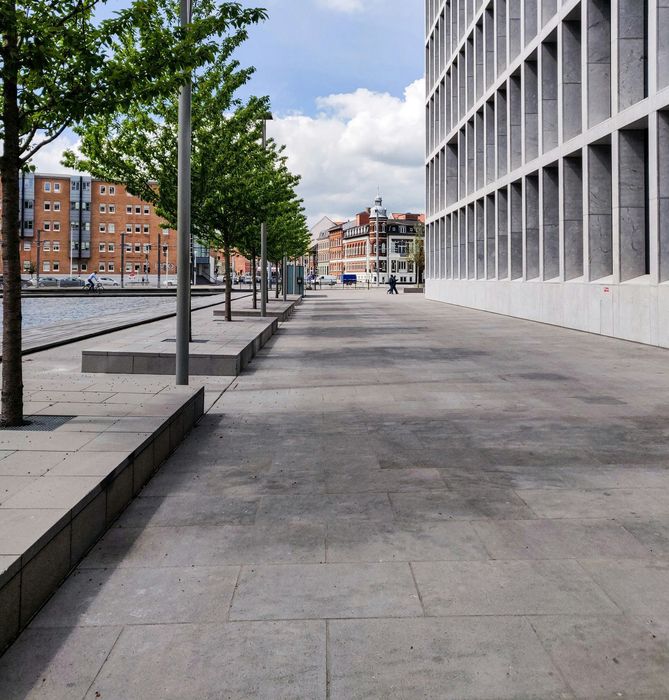 Empty urban walkway beside a modern white building, with trees and apartment blocks in the distance.