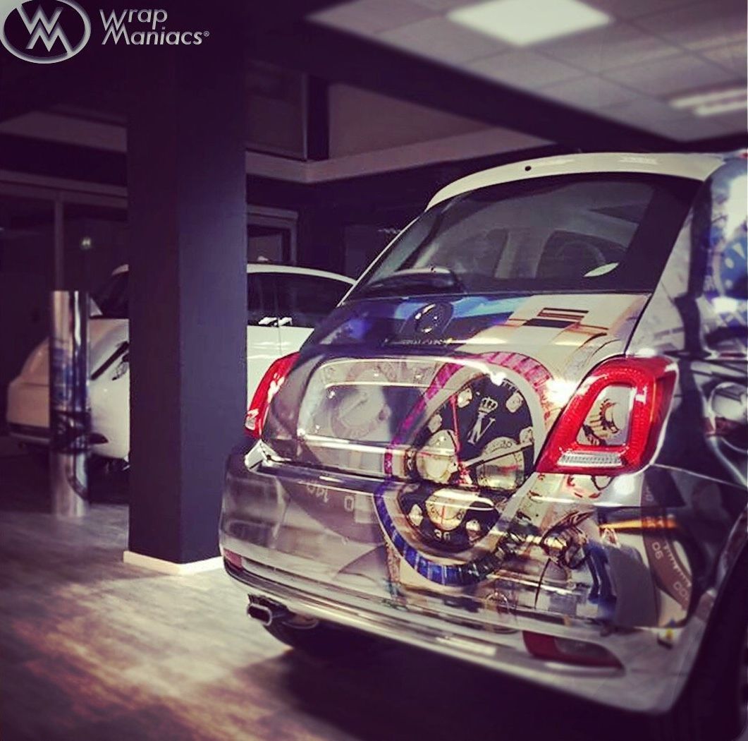 Chrome-wrapped Fiat 500 with a complex graphic of a Rolex on the rear, parked inside a shop with other vehicles.
