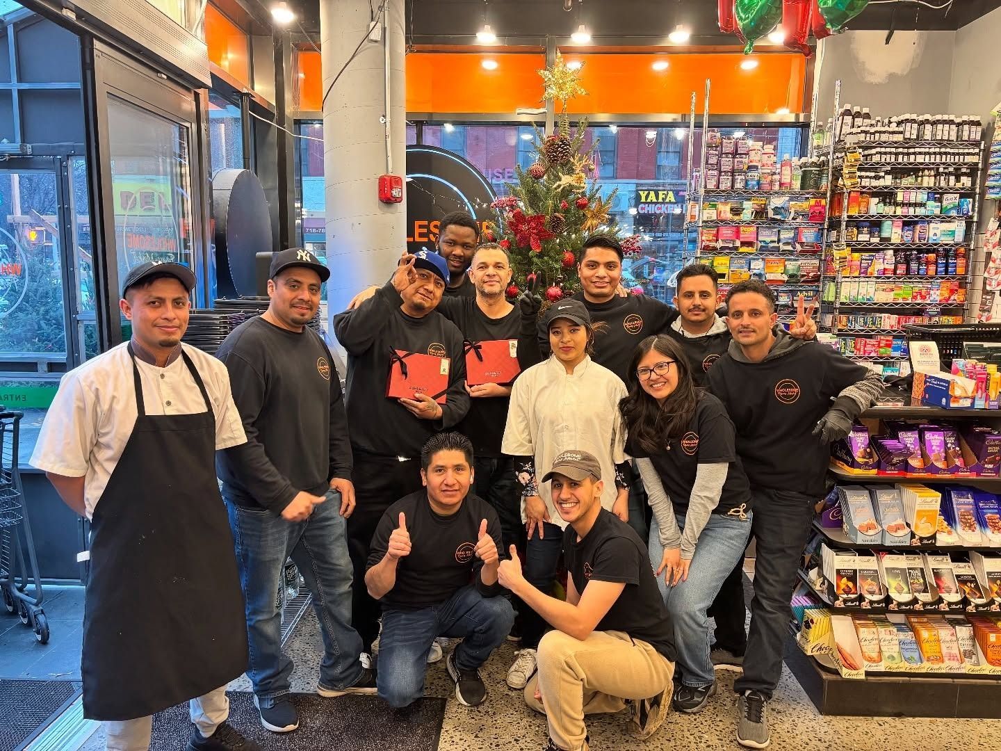 Group of people posing inside a brightly lit convenience store, with shelves, snacks, and a small Christmas tree.