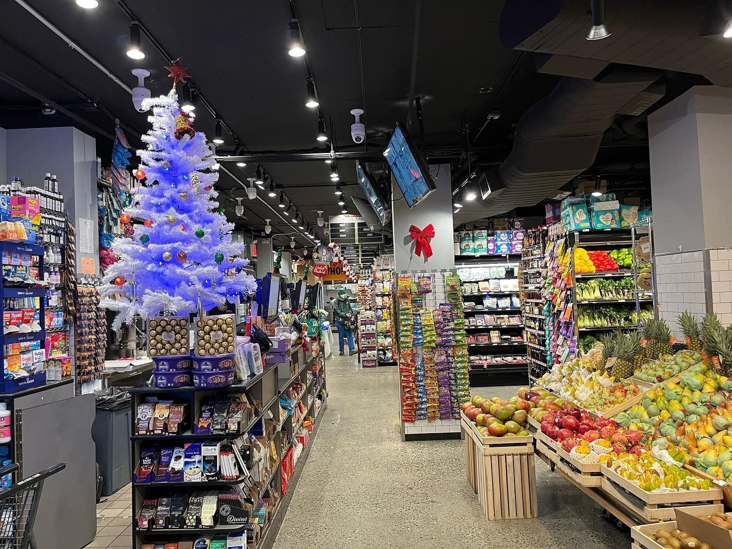 Grocery store aisle with a lit blue Christmas tree and produce displays under bright ceiling lights