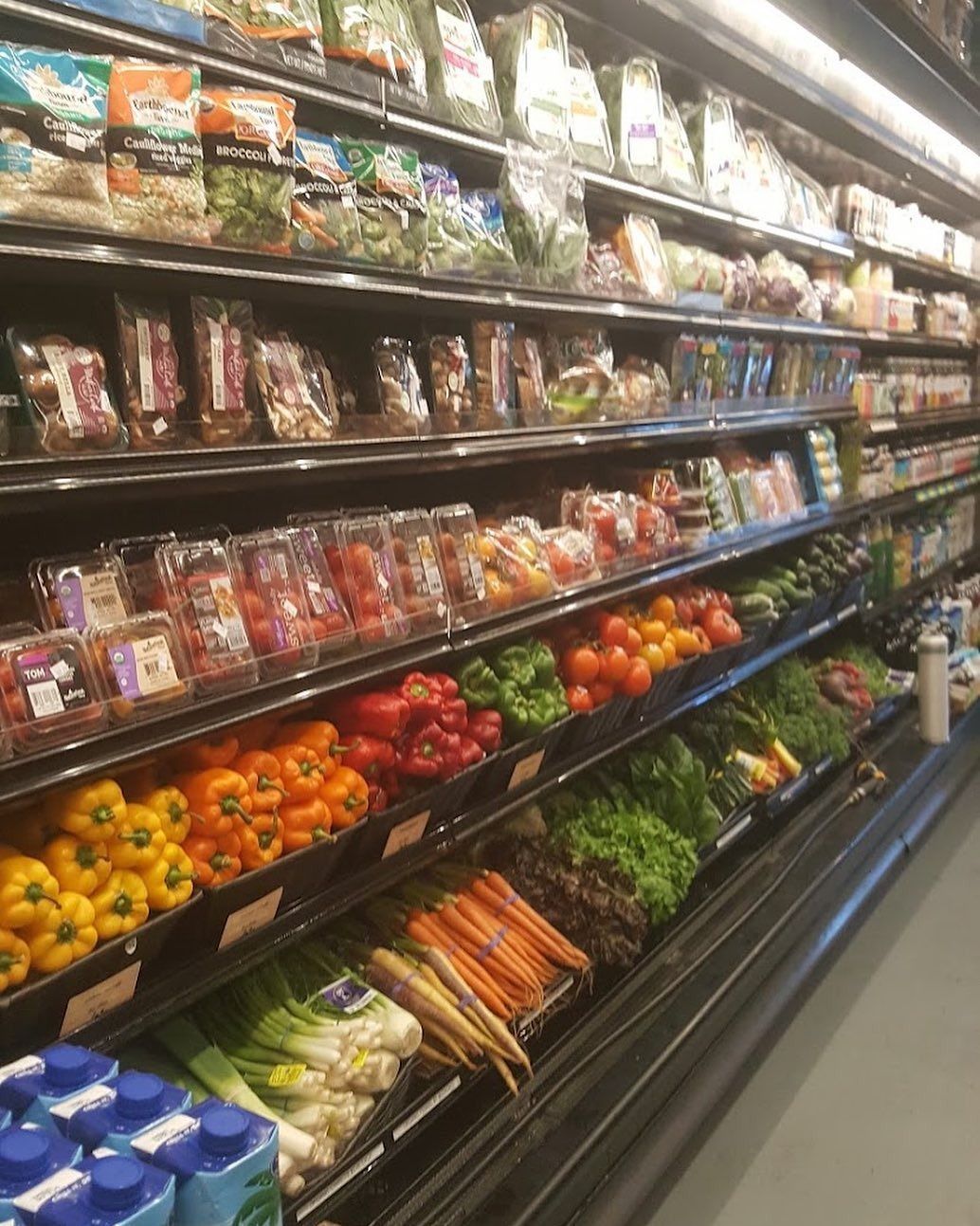 Grocery store produce aisle with shelves of packaged greens, carrots, tomatoes, and oranges.