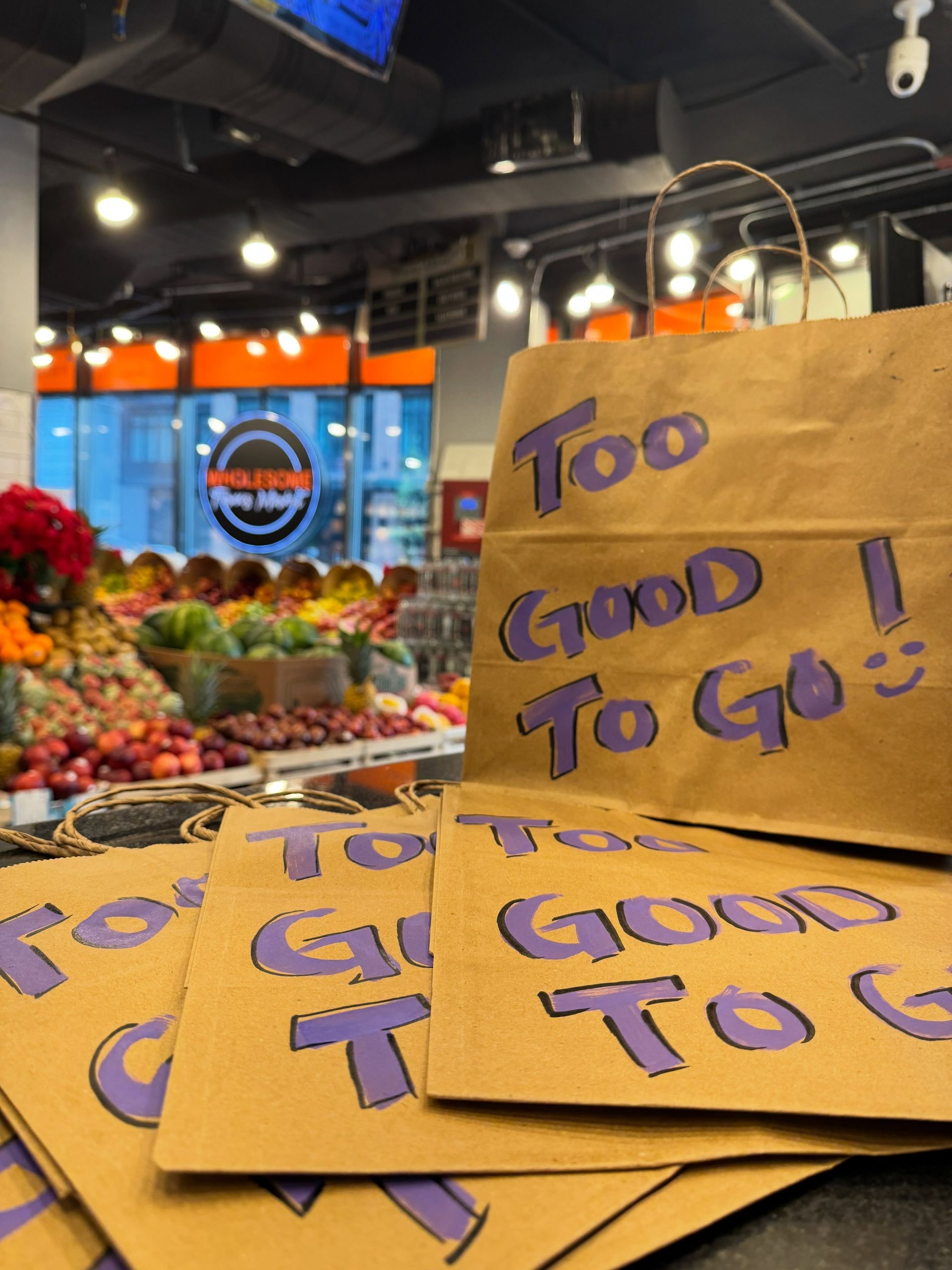 Handwritten “Too Good To Go” signs at a grocery store counter with produce in the background