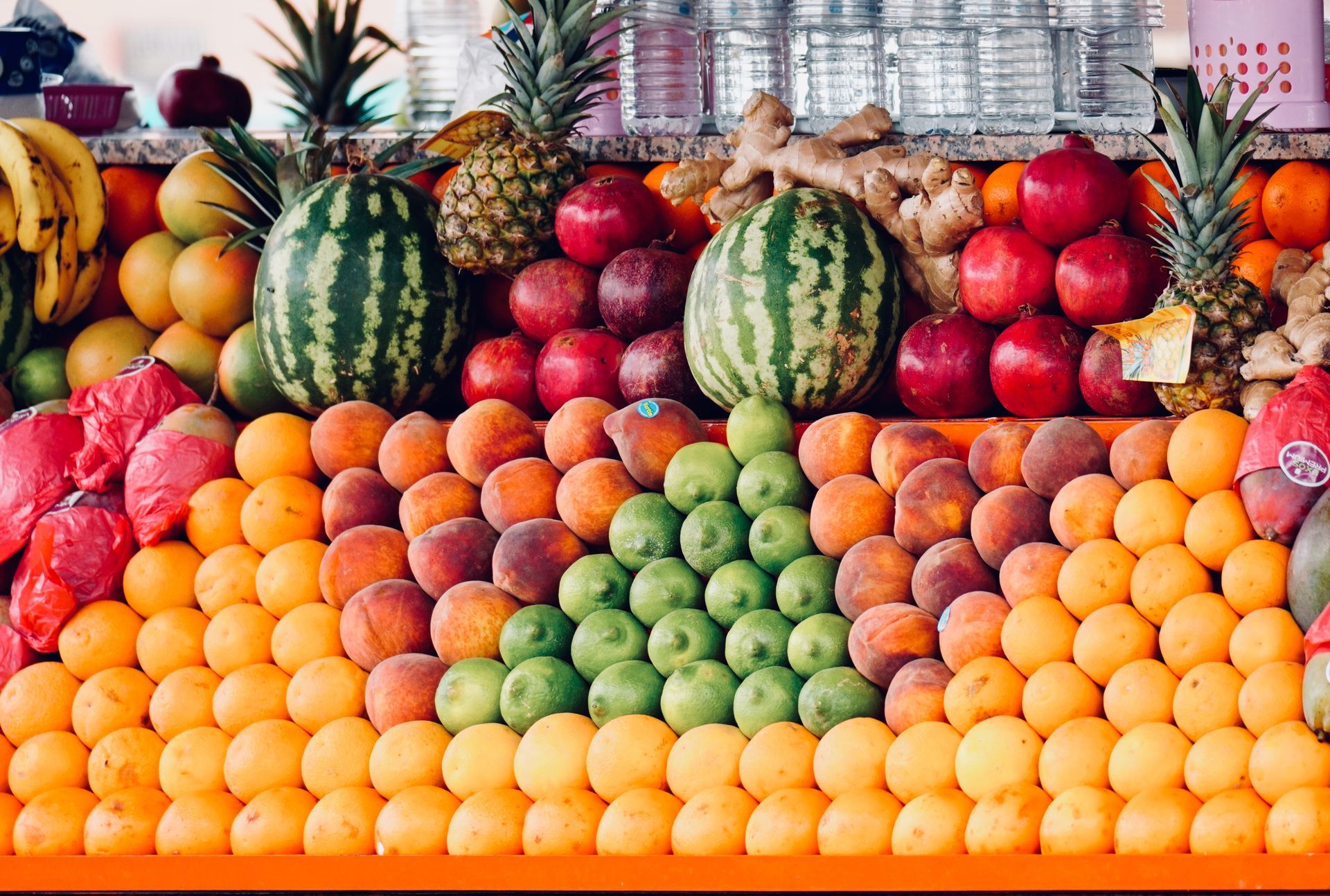 Colorful fruit display with oranges, apples, watermelons, grapes, and pineapples stacked in a market stall