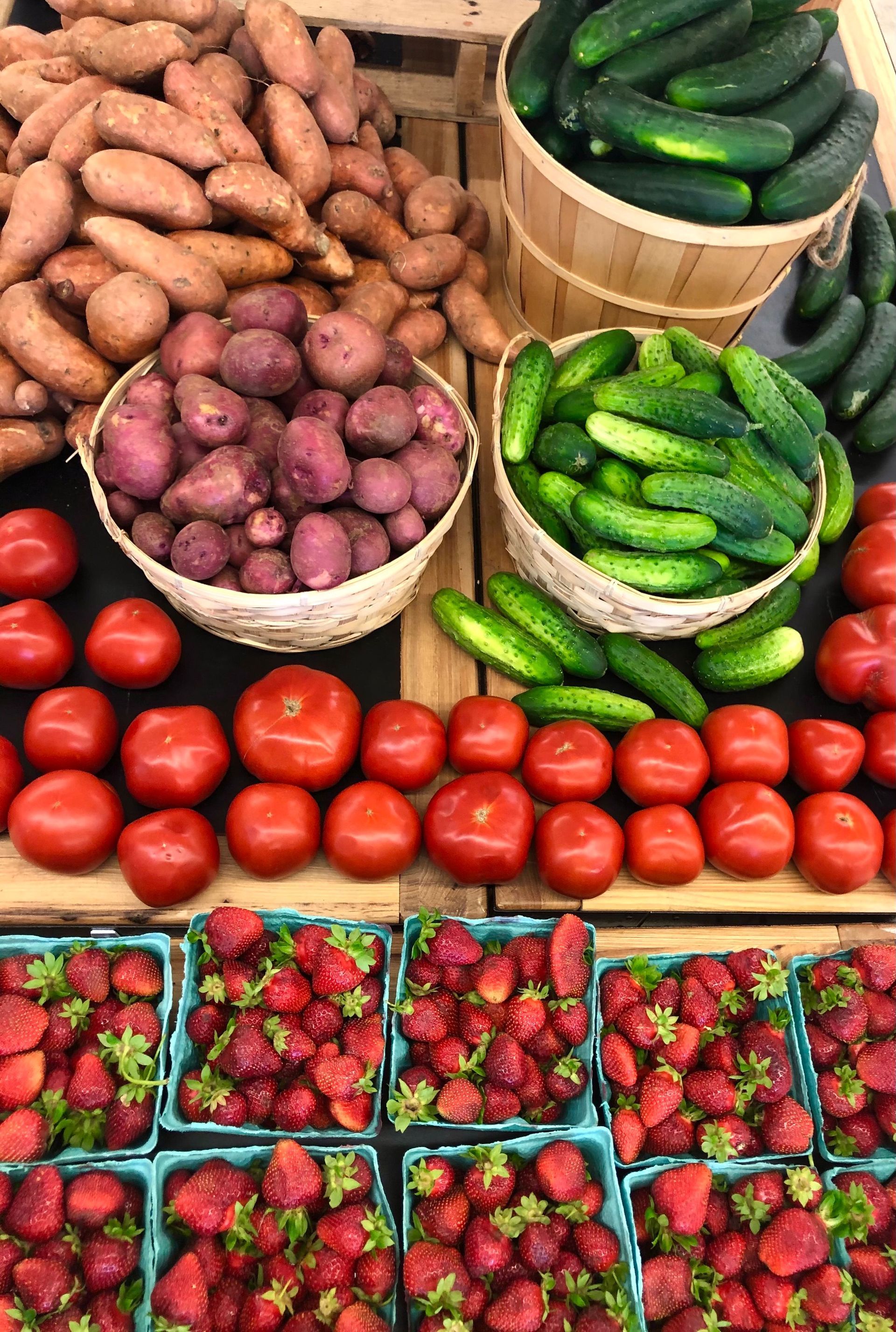 Market stall with baskets of sweet potatoes and cucumbers, rows of tomatoes, and trays of strawberries.