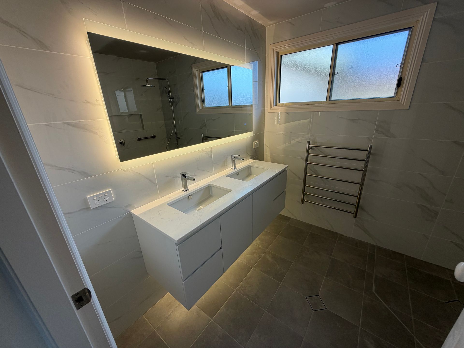 Modern bathroom with lit mirror, white vanity, dark tile floor, and towel rack by window.