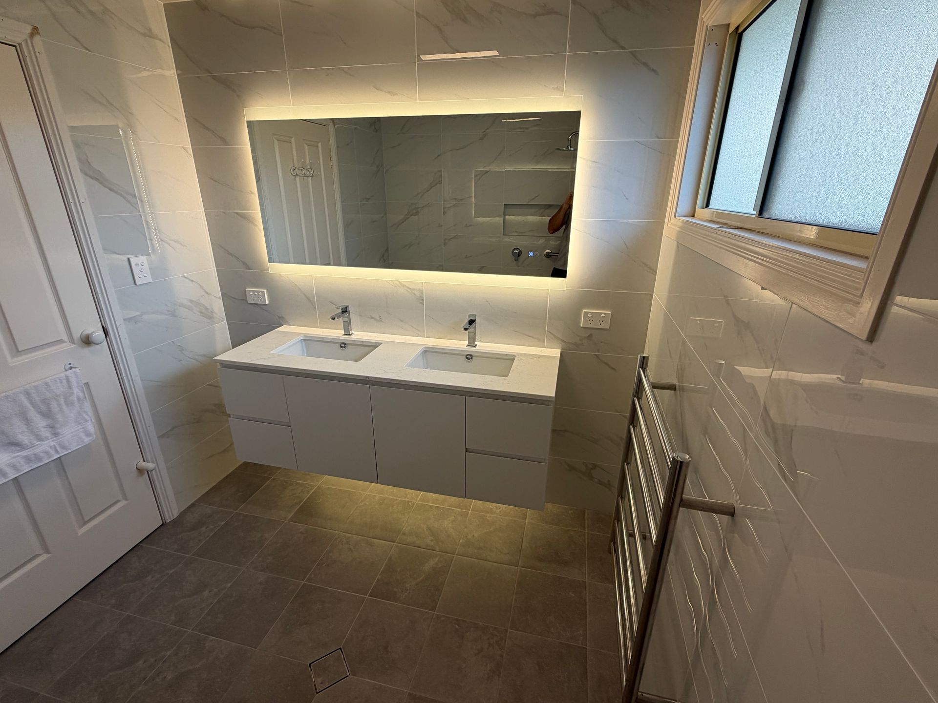 Modern bathroom with double sink vanity, large backlit mirror, white tile walls, and a towel radiator