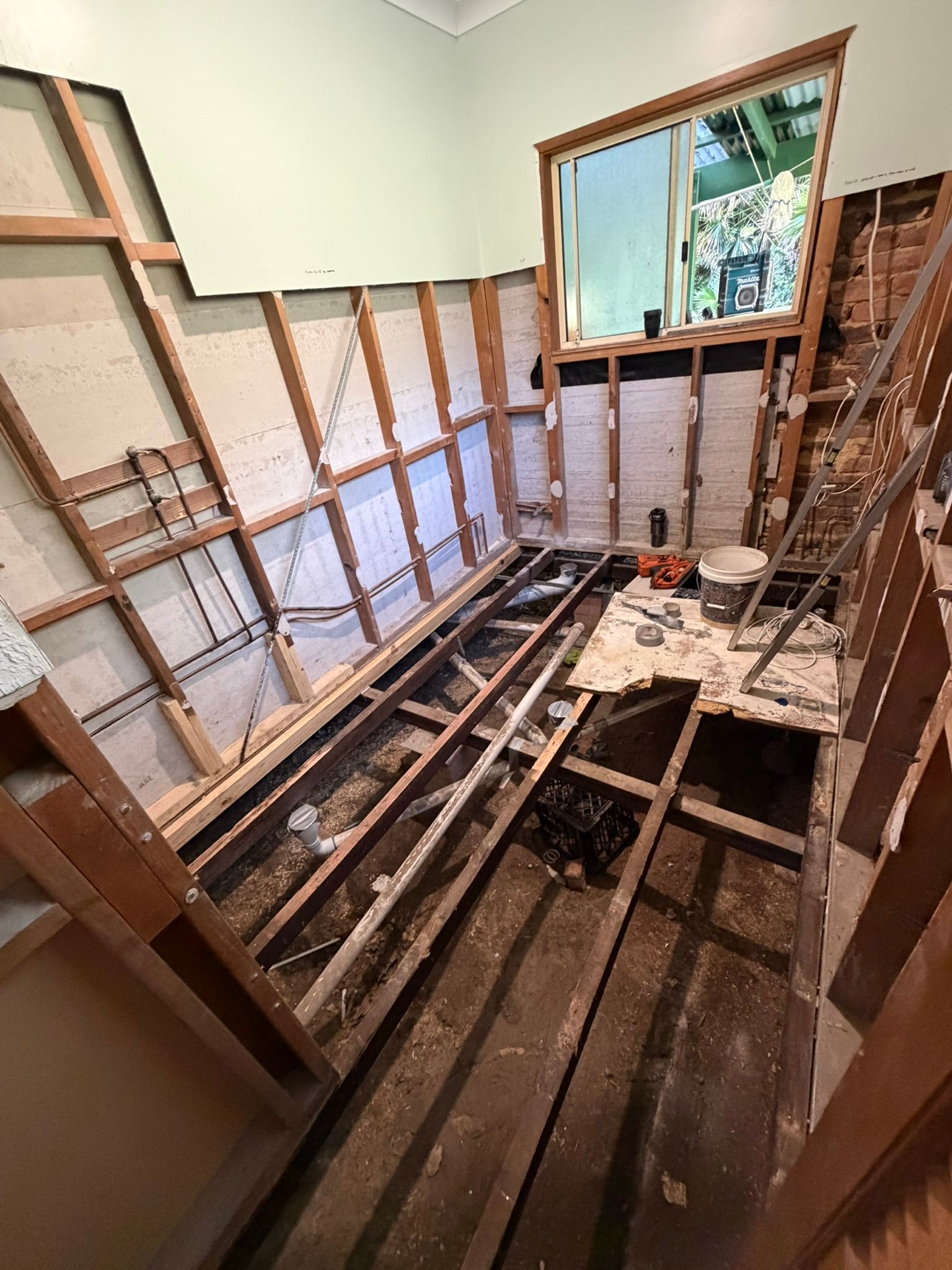 Partially gutted room with exposed floor joists and wall framing, window at end, tools on debris-covered floor