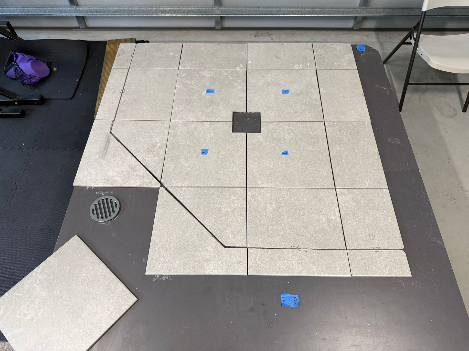 Gray floor with taped rectangular layout, blue markers, and a small vent; one loose tile at lower left.