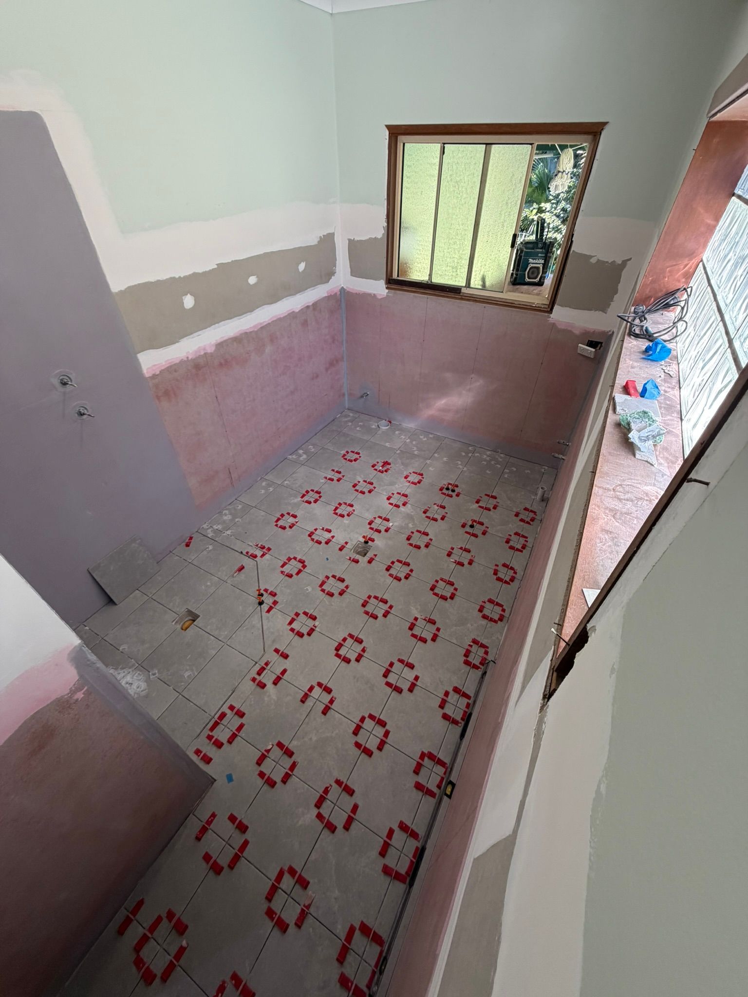 Empty unfinished bathroom with pink and white tiled walls, a window, and patterned floor tiles.