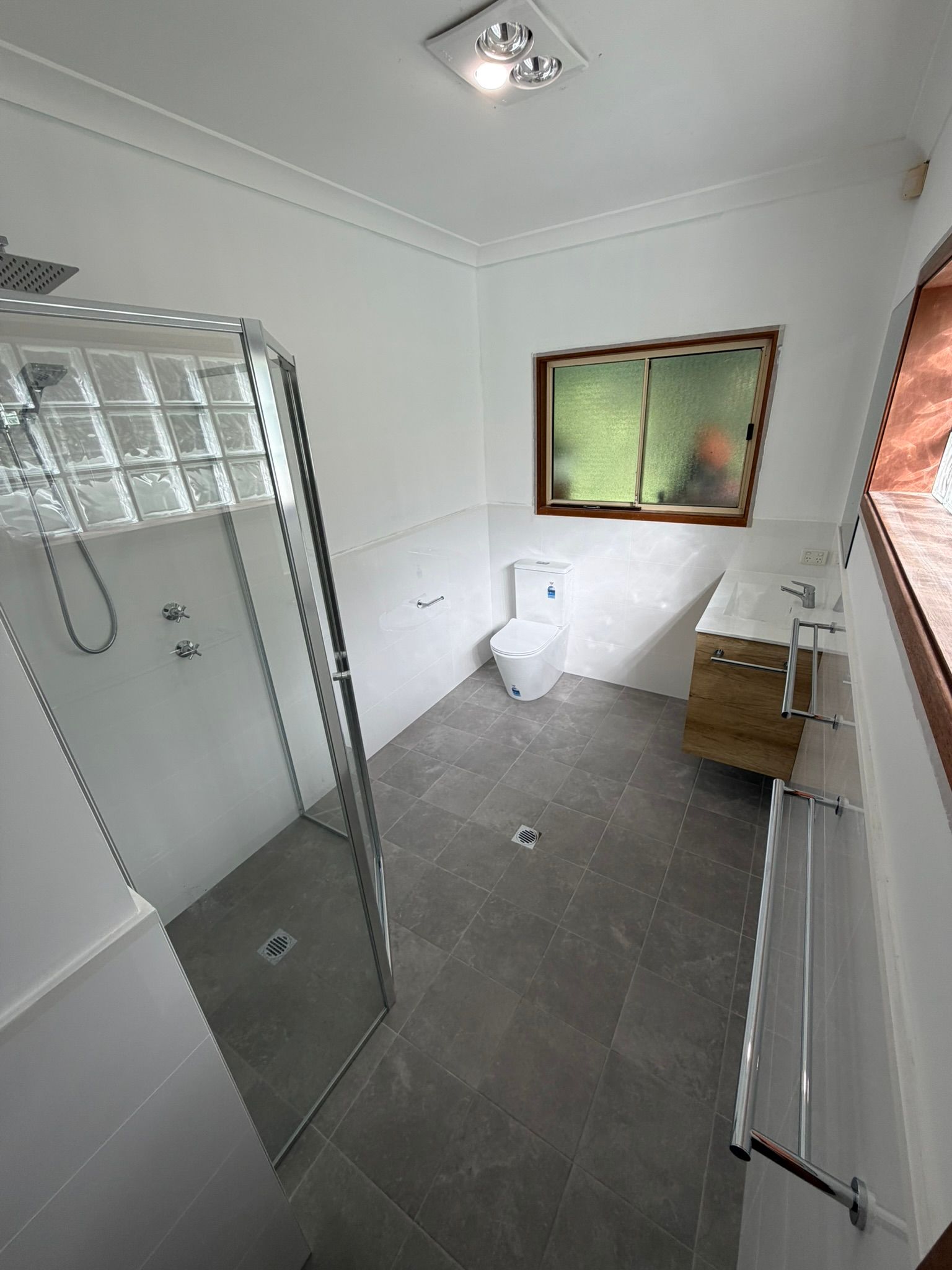 Small white tiled bathroom with shower, toilet, sink, mirror, and window blinds.