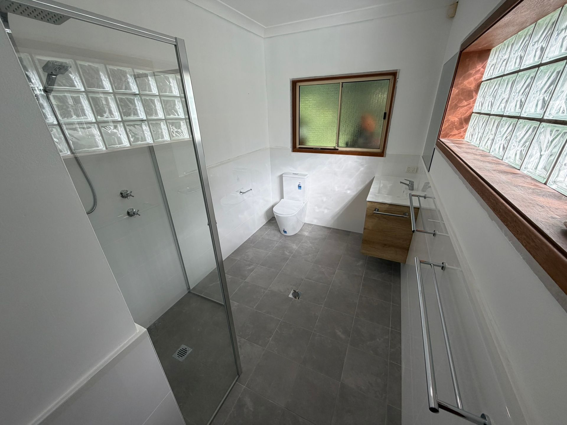 Small bathroom with glass shower, toilet, gray tile floor, vanity, and frosted windows.