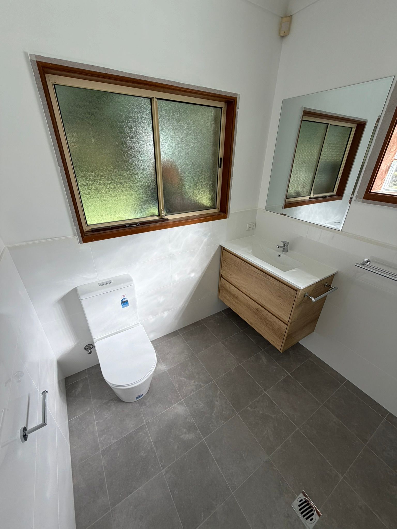 Small bathroom with toilet, floating vanity, mirror, frosted window, and gray tile floor.