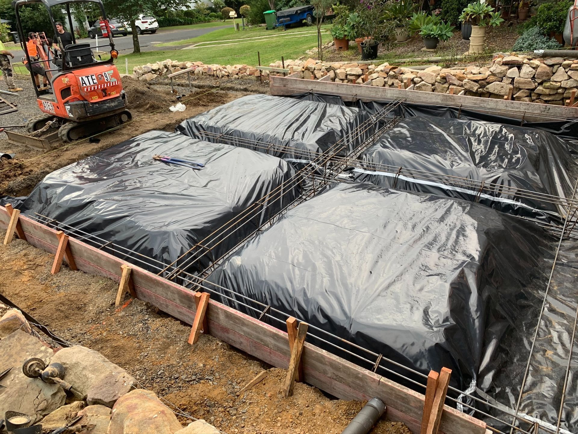 Excavator digging near a lined rectangular pit covered with black plastic on a construction site