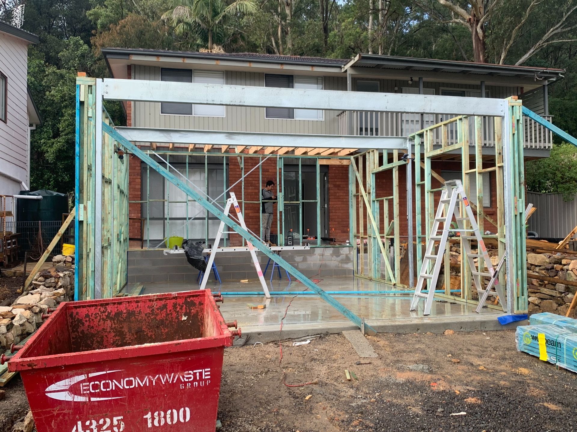 House under construction with exposed framing, scaffolding, and a red debris dumpster in front