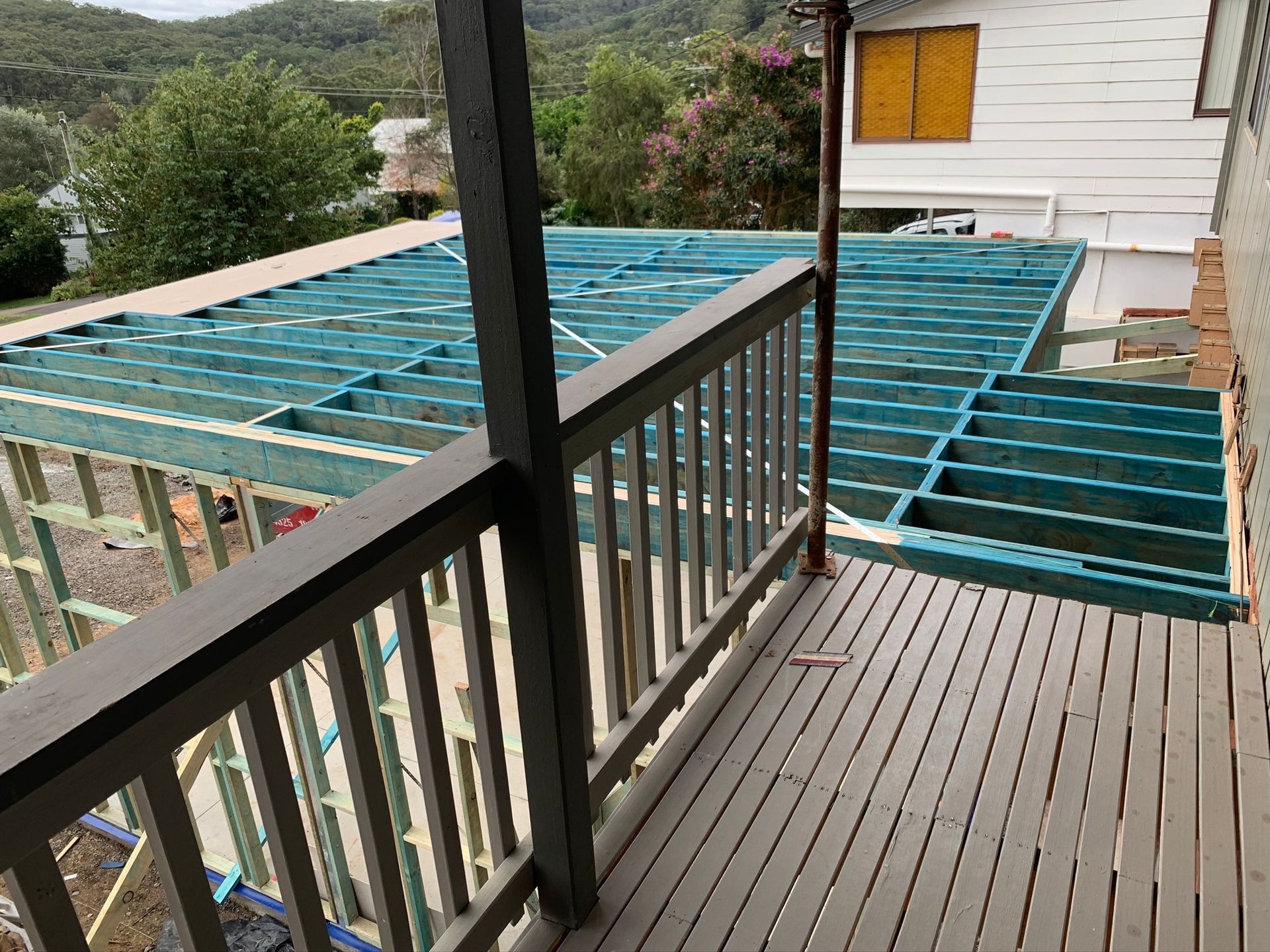 Deck under construction with exposed blue framing beside a railing and house exterior.