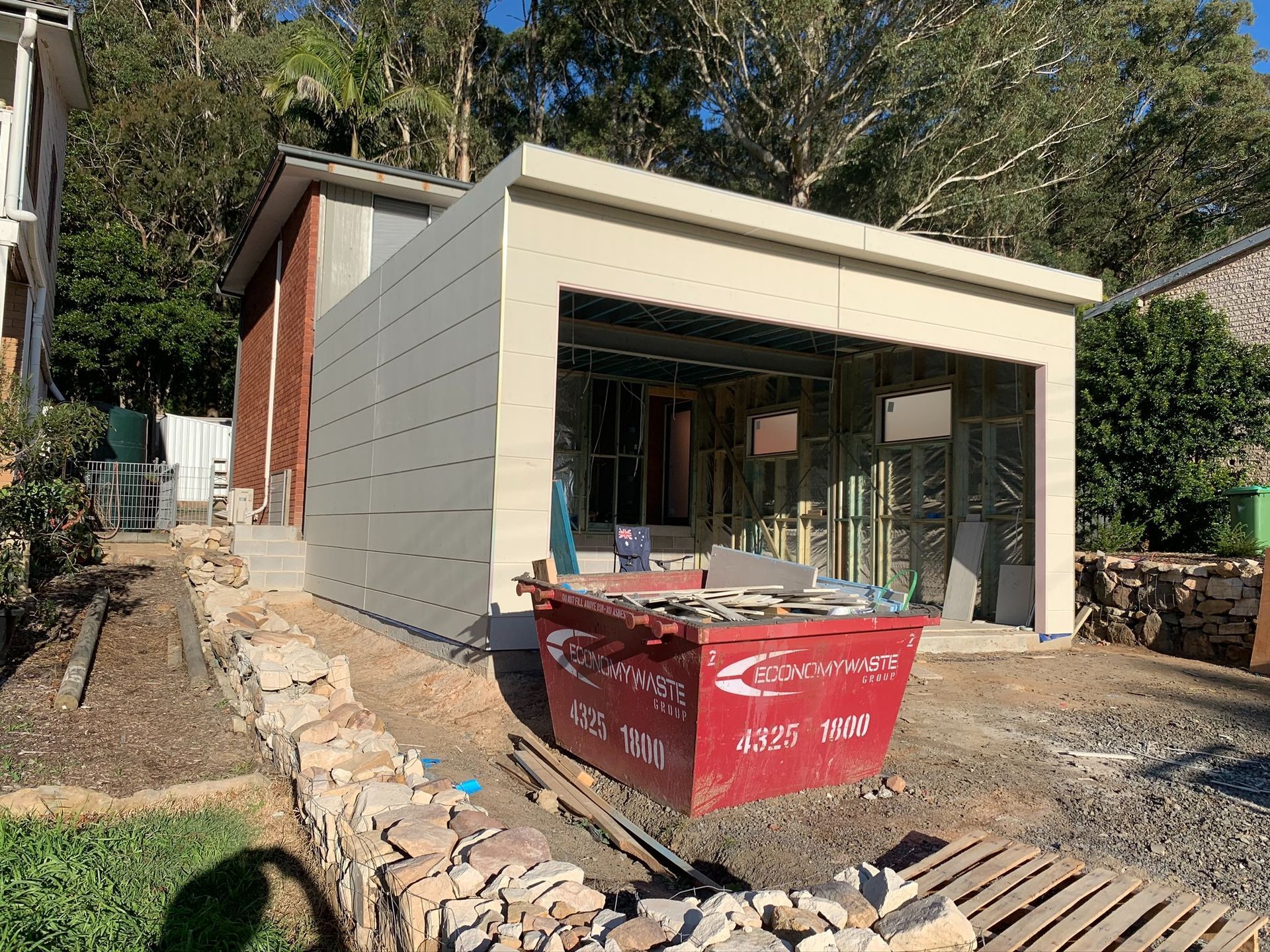 Modern house under construction with red dumpster in front and stone retaining wall along the driveway