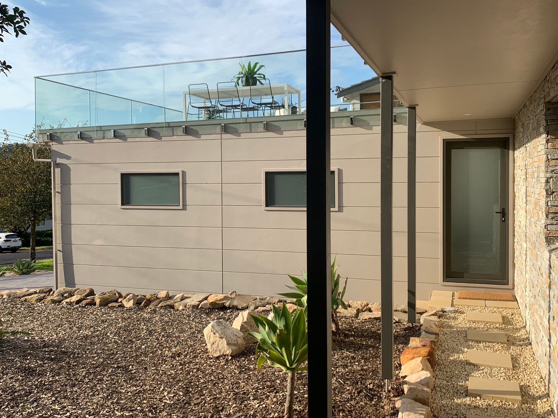 Modern stucco house exterior with small windows, glass rooftop railing, and stone walkway under a covered patio