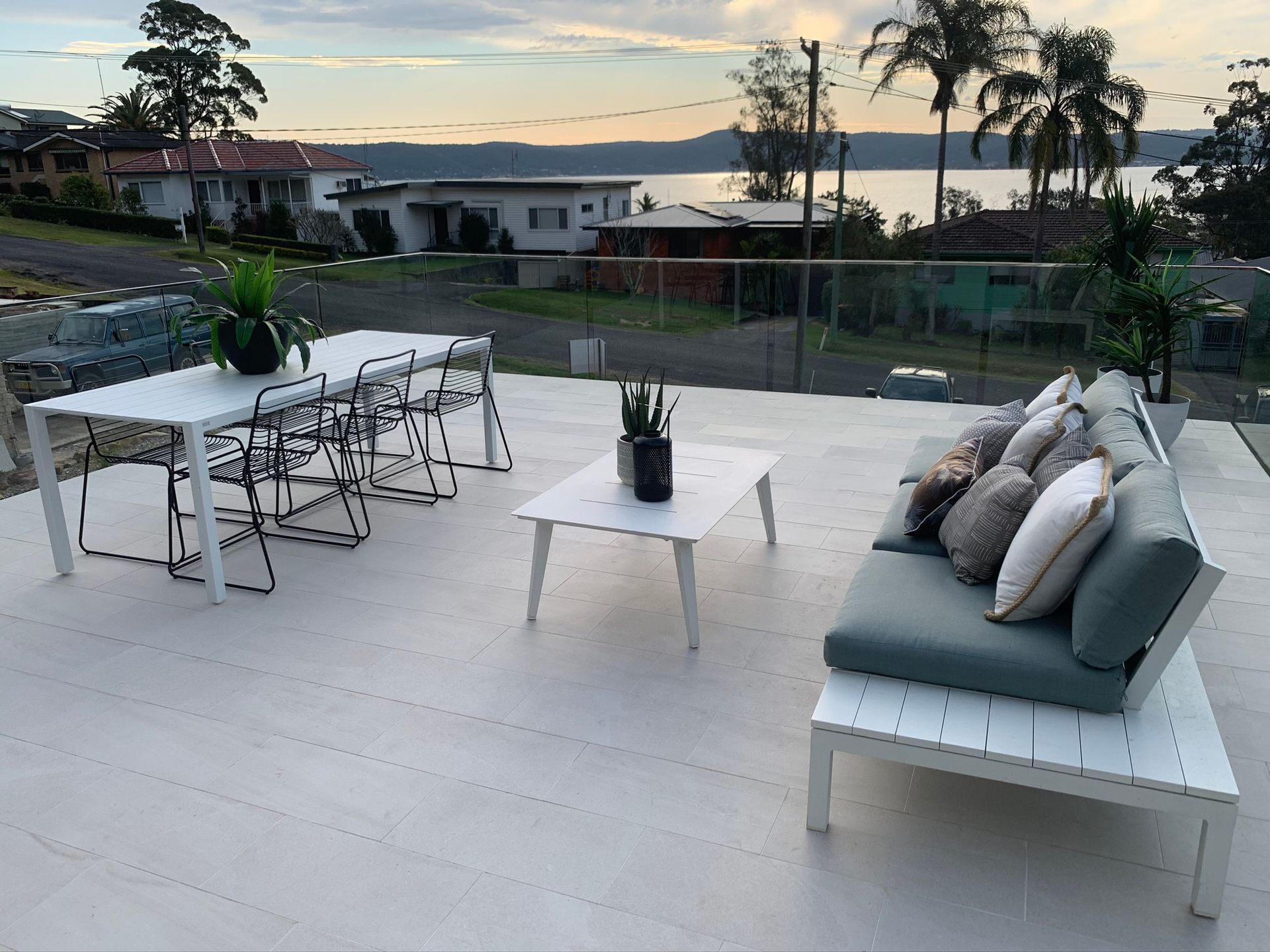 Patio with white table and chairs, cushioned bench, potted plants, and sunset view over water