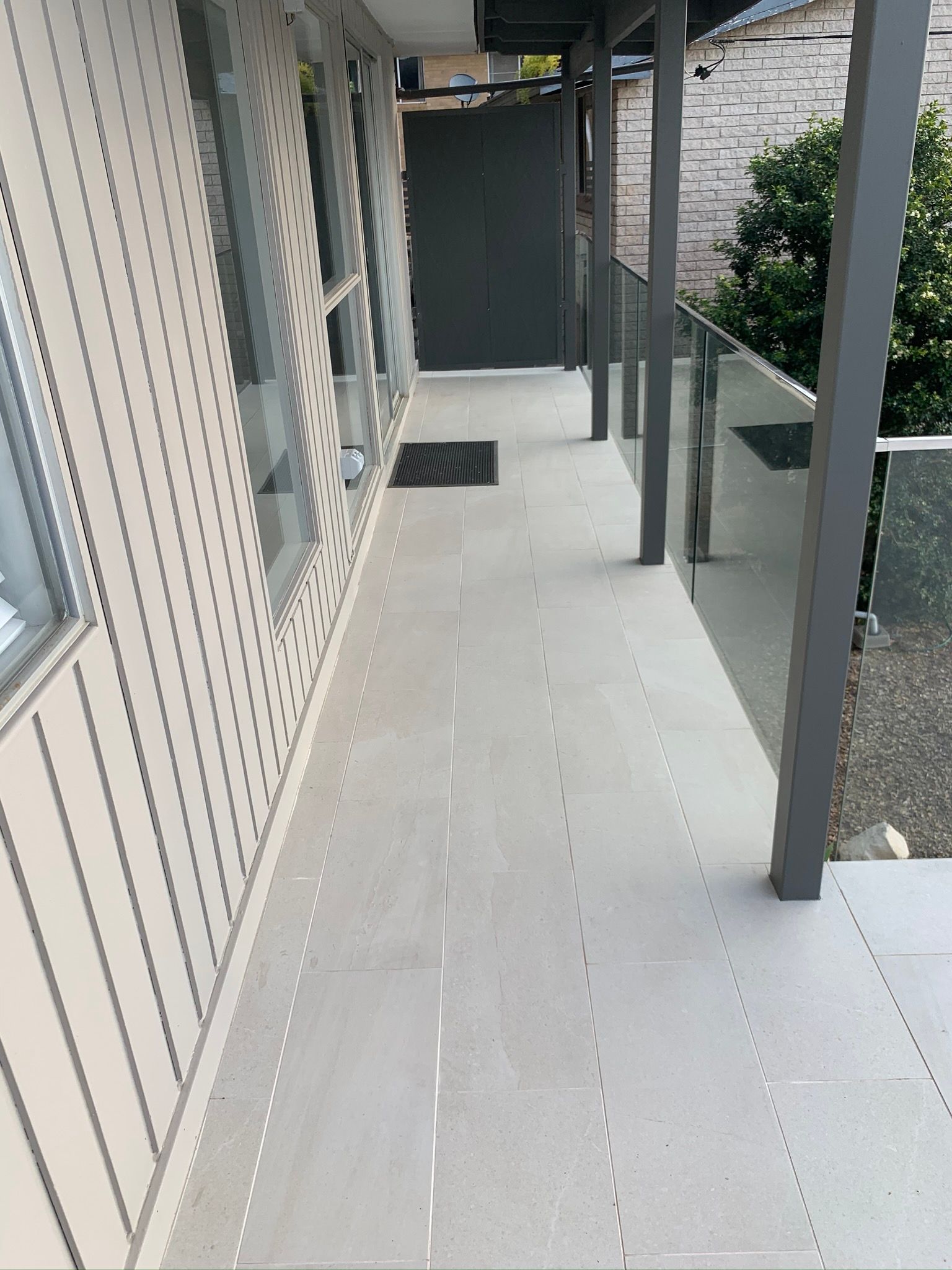 Narrow apartment balcony with light flooring, gray railings, and a dark door at the end.