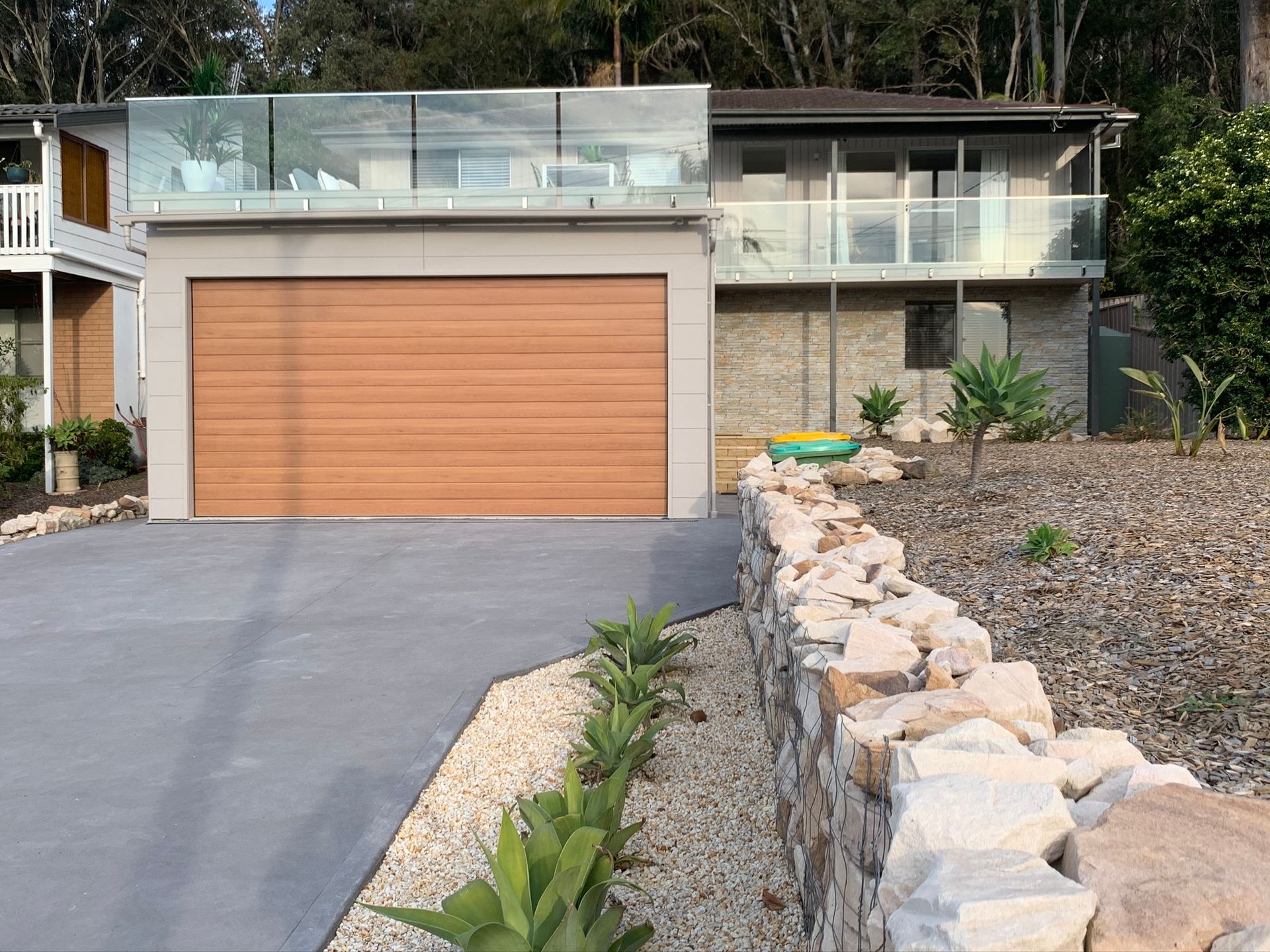 Modern house with wood garage door, glass balcony, and sloped driveway beside a rock retaining wall