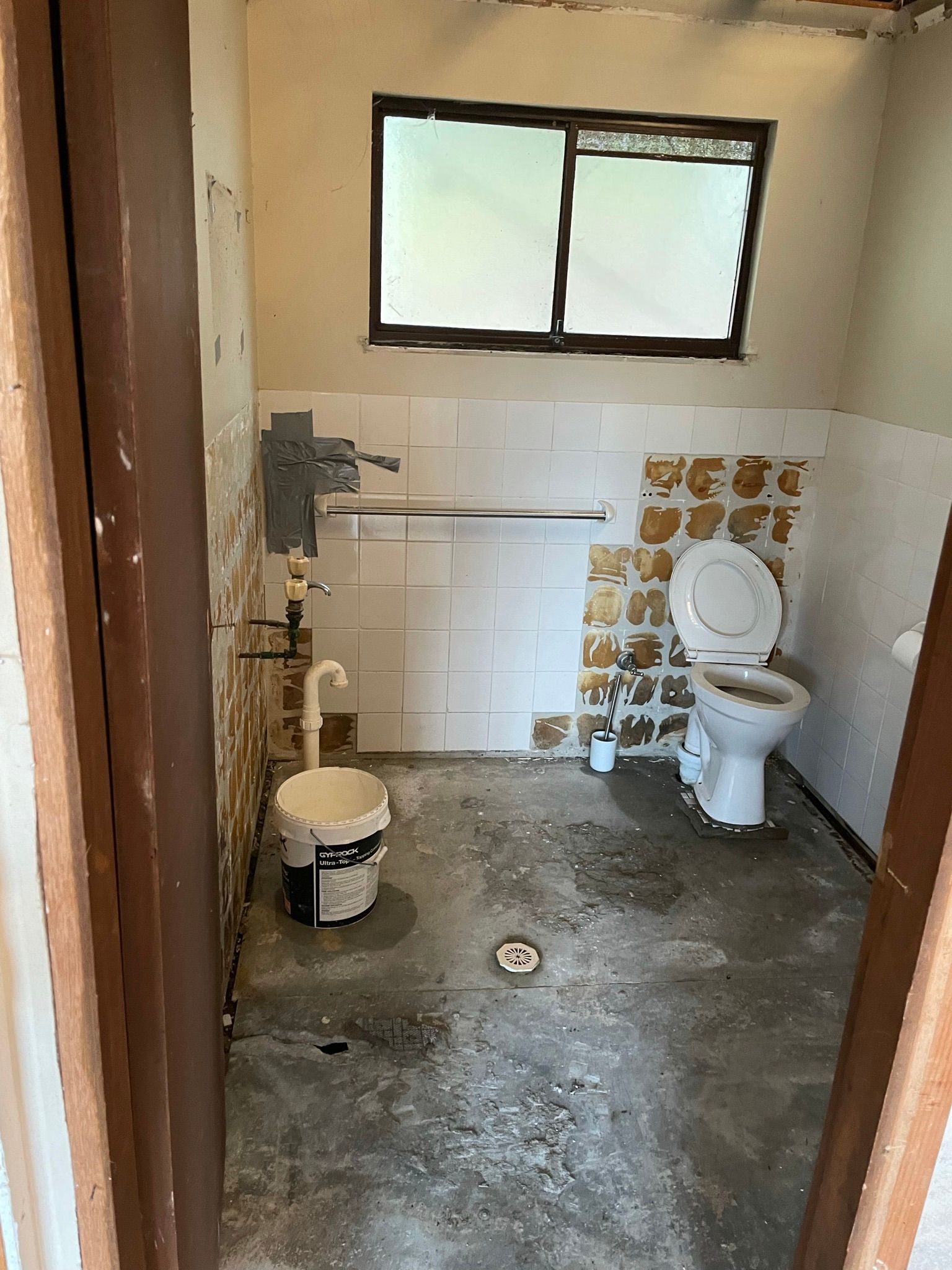 Small unfinished bathroom with toilet, window, exposed plumbing, and a concrete floor.