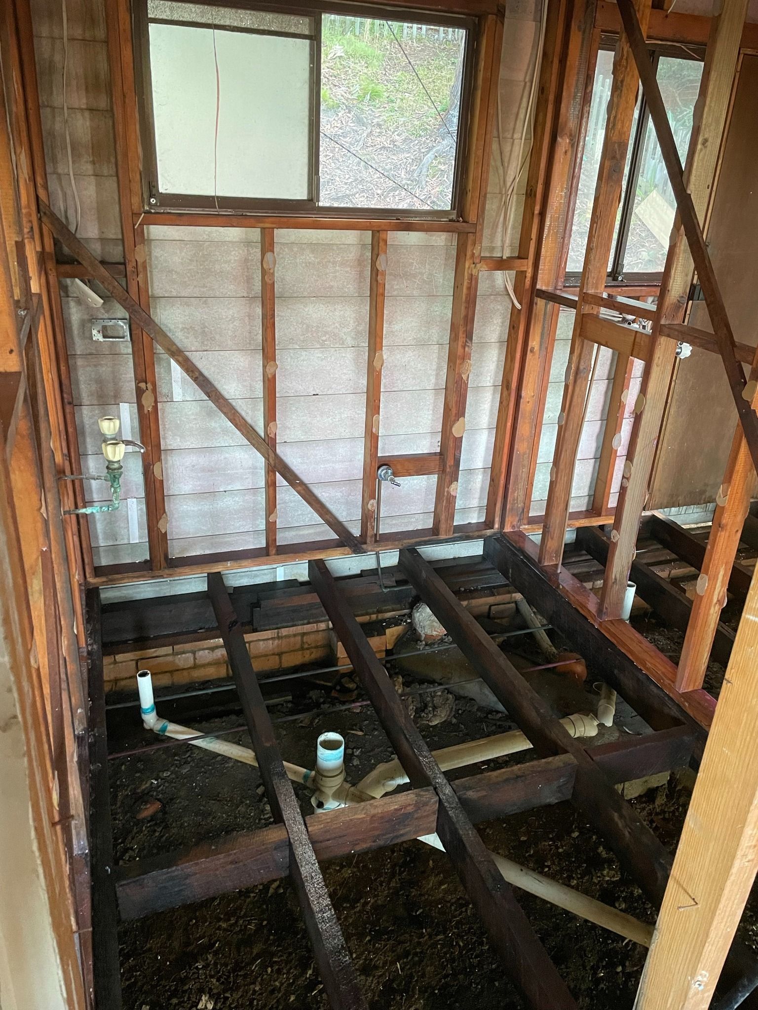 Interior wall under renovation with exposed studs, floor joists, and a window covered by plastic sheeting.