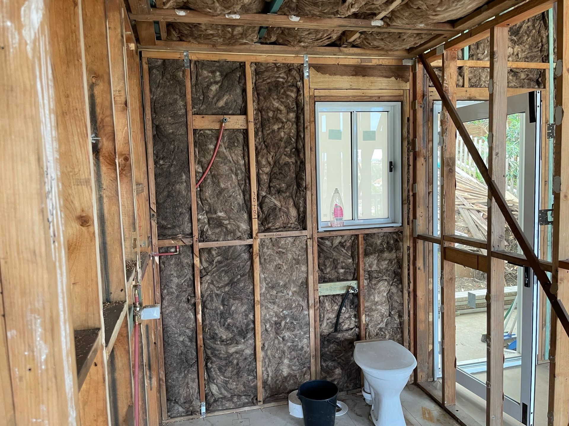Partially demolished bathroom with exposed studs, window, toilet, and bucket on the floor.