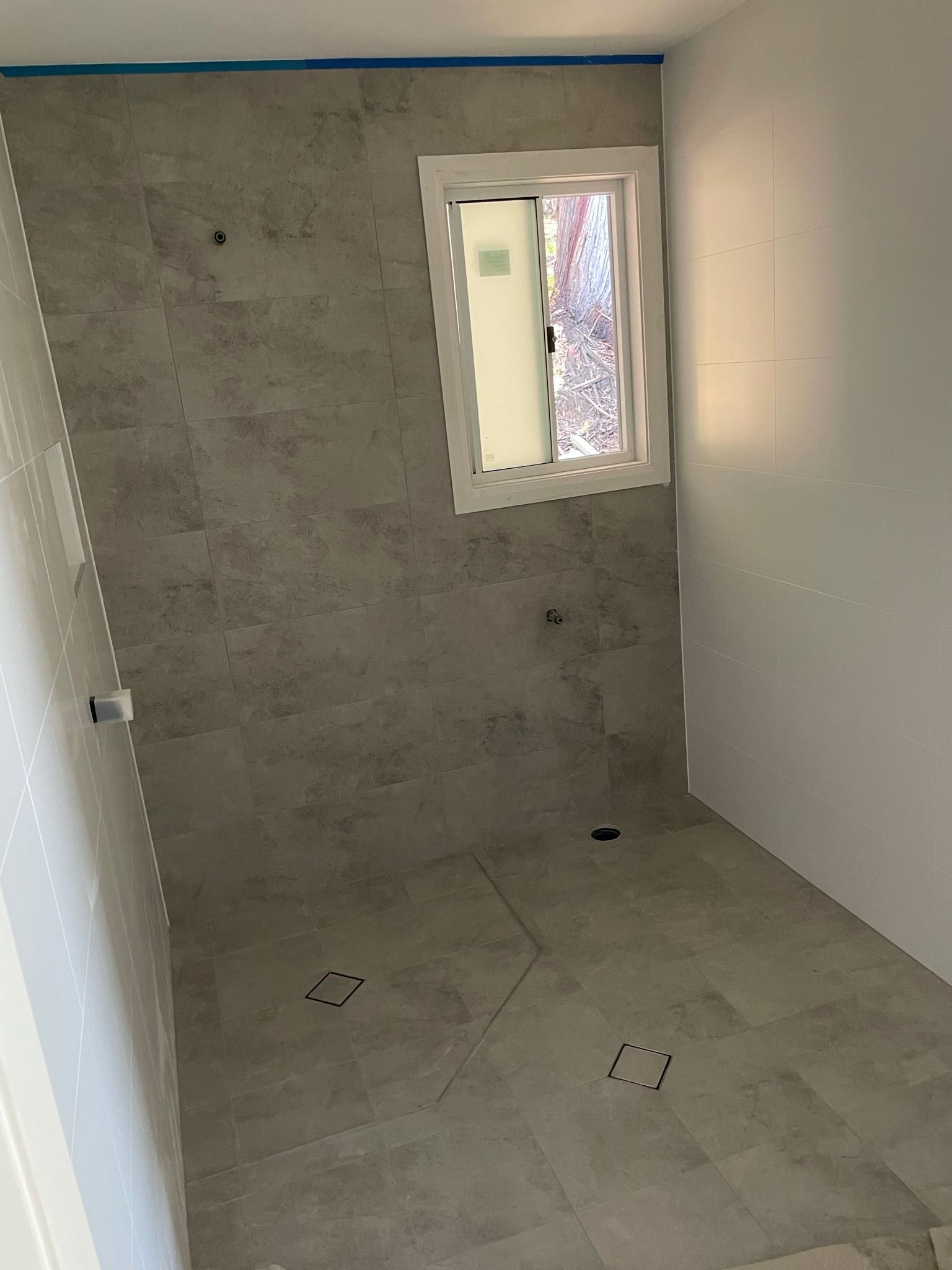 Empty unfinished room with gray tiled walls and floor, white wall, and a small window.
