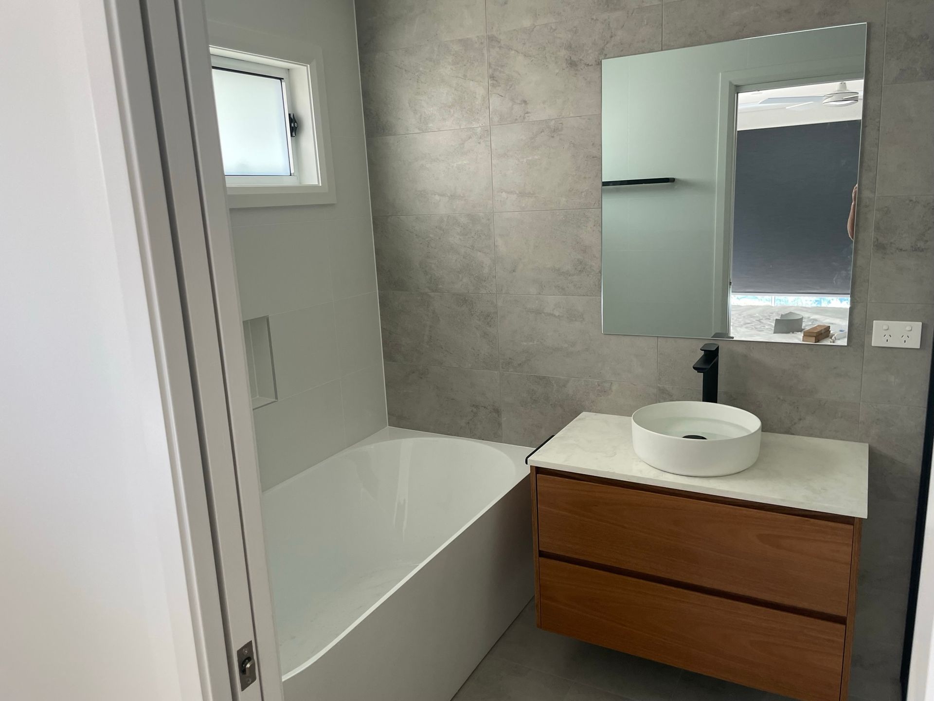 Modern bathroom with white tub, gray tile walls, and a wooden vanity with round sink and mirror.