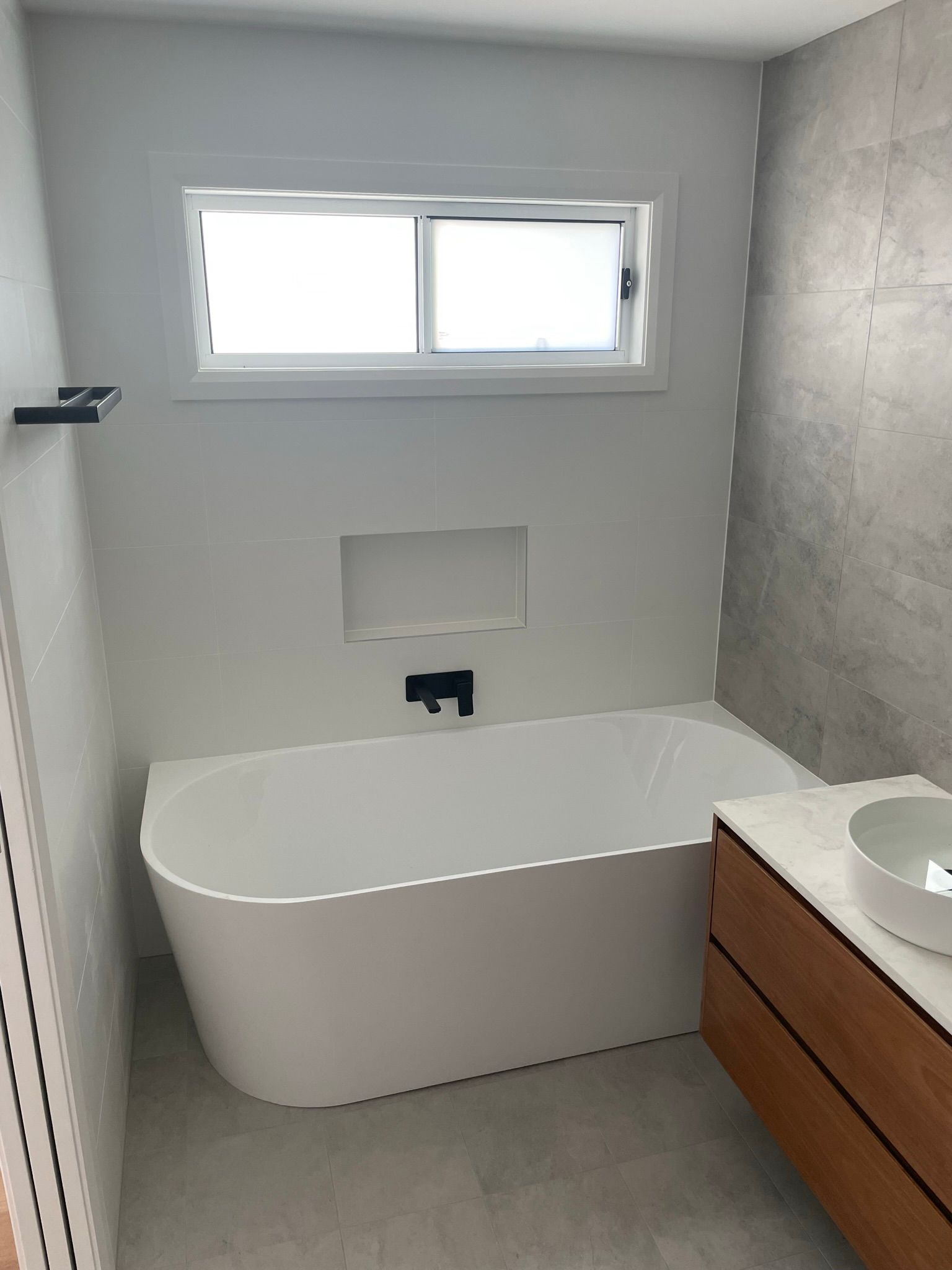 Modern bathroom with a freestanding tub, window above, and wood vanity beside a concrete shower wall