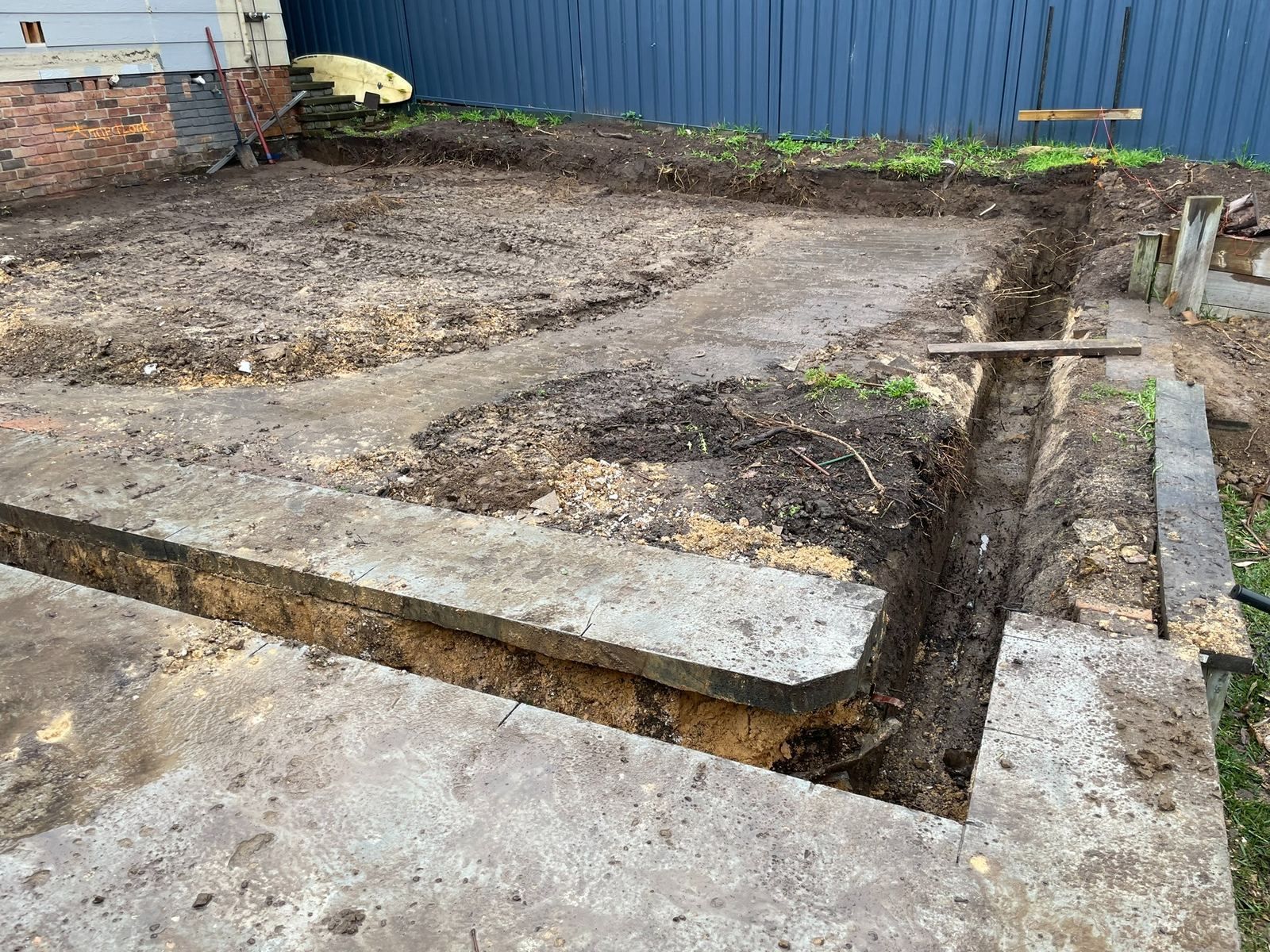 Excavated backyard foundation area with concrete footings, trenches, and a blue fence in the background