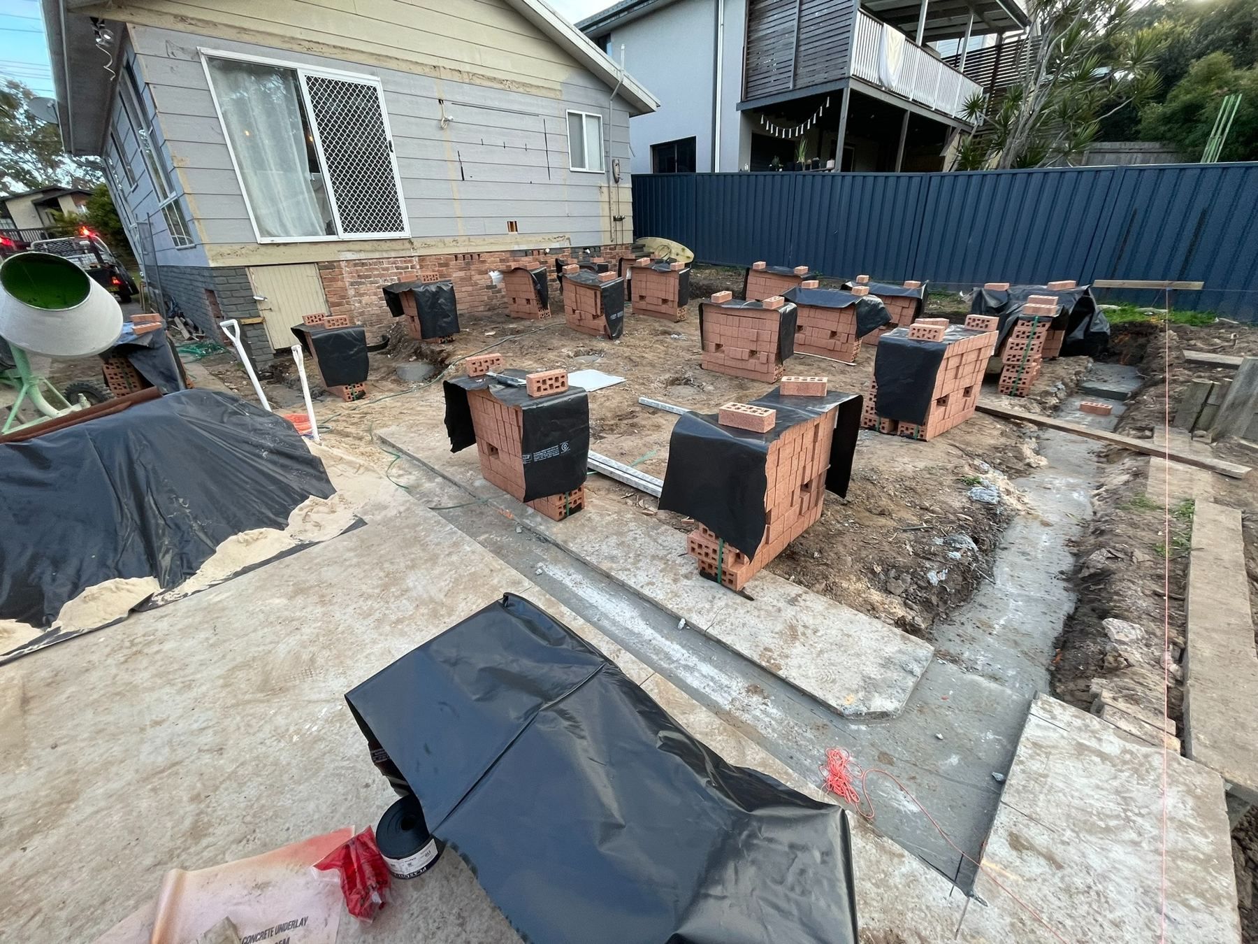 Backyard construction area with brick piers, concrete slabs, and a house beside a blue fence
