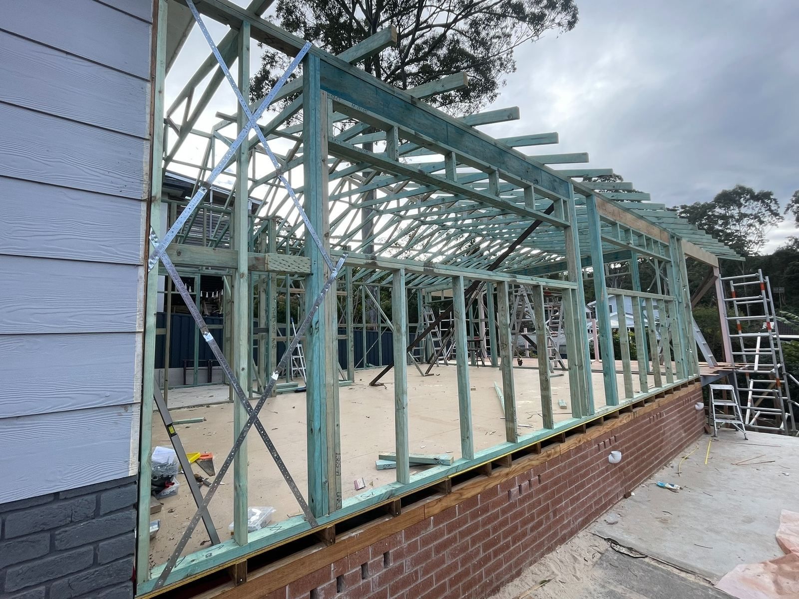 Framed house extension under construction beside a brick wall, with exposed timber studs and scaffolding.