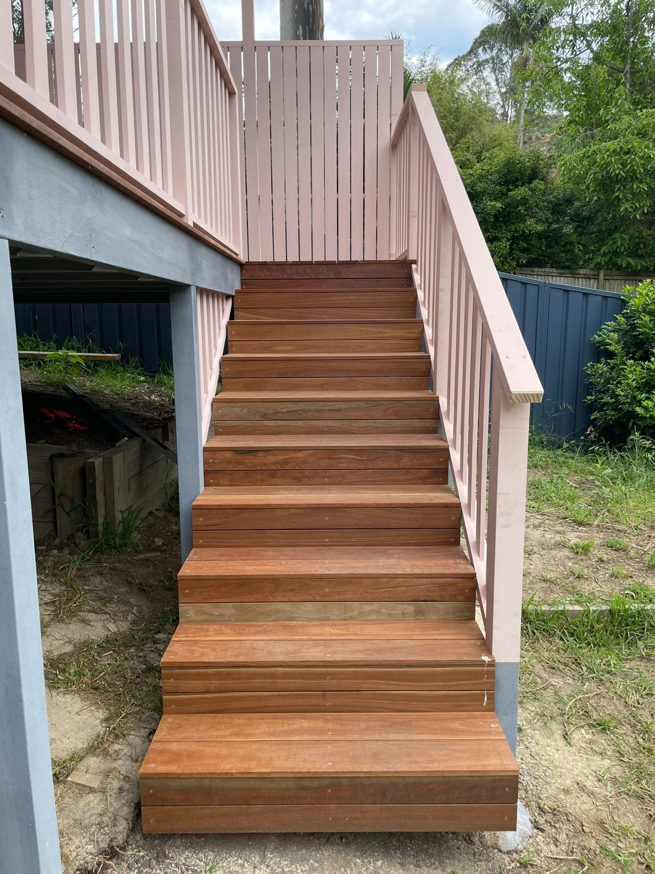 Wooden outdoor staircase with pale pink railings leading to a raised deck in a backyard.