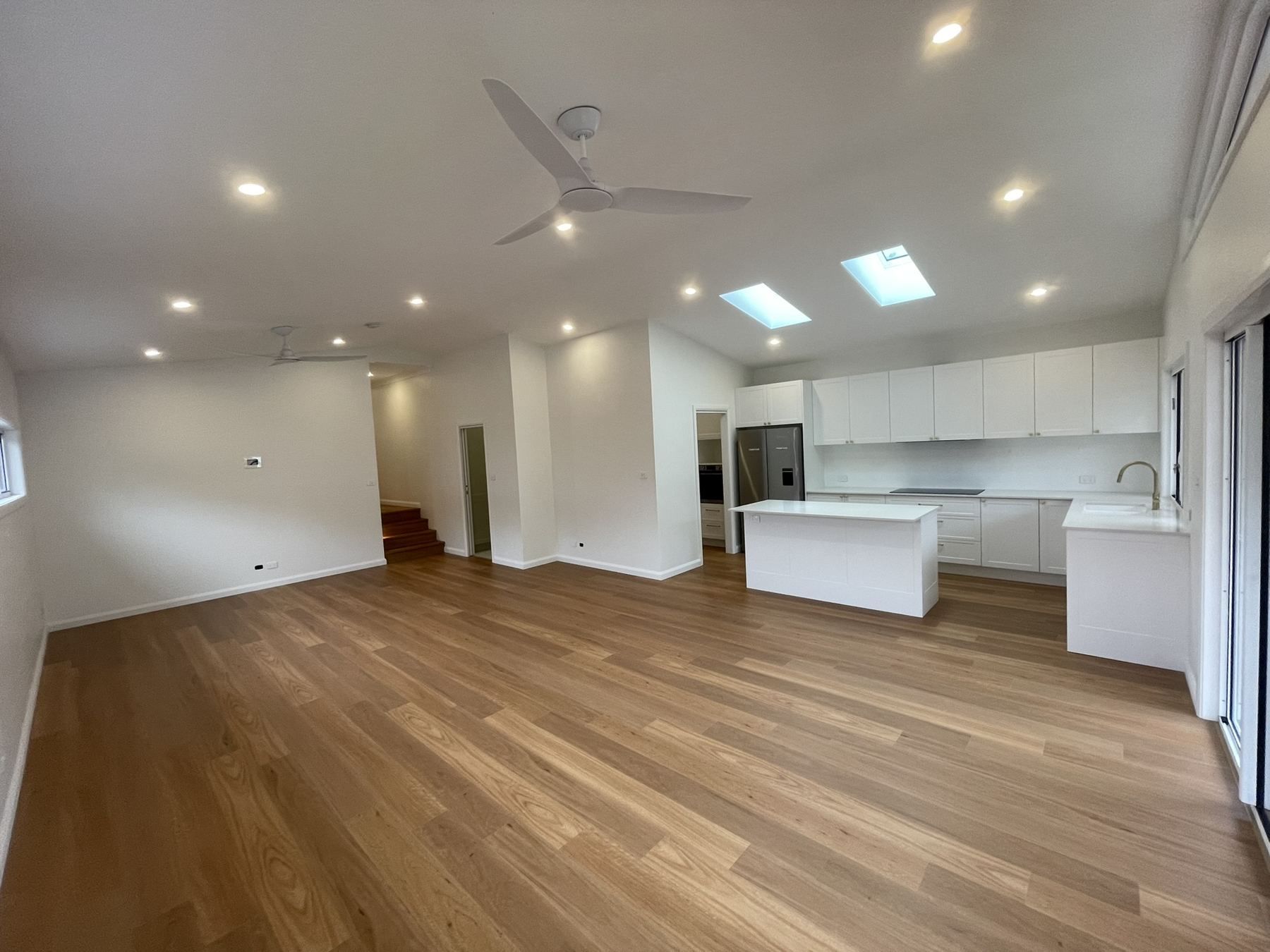Spacious empty modern living room with wood floors, white walls, recessed lights, and an open kitchen.