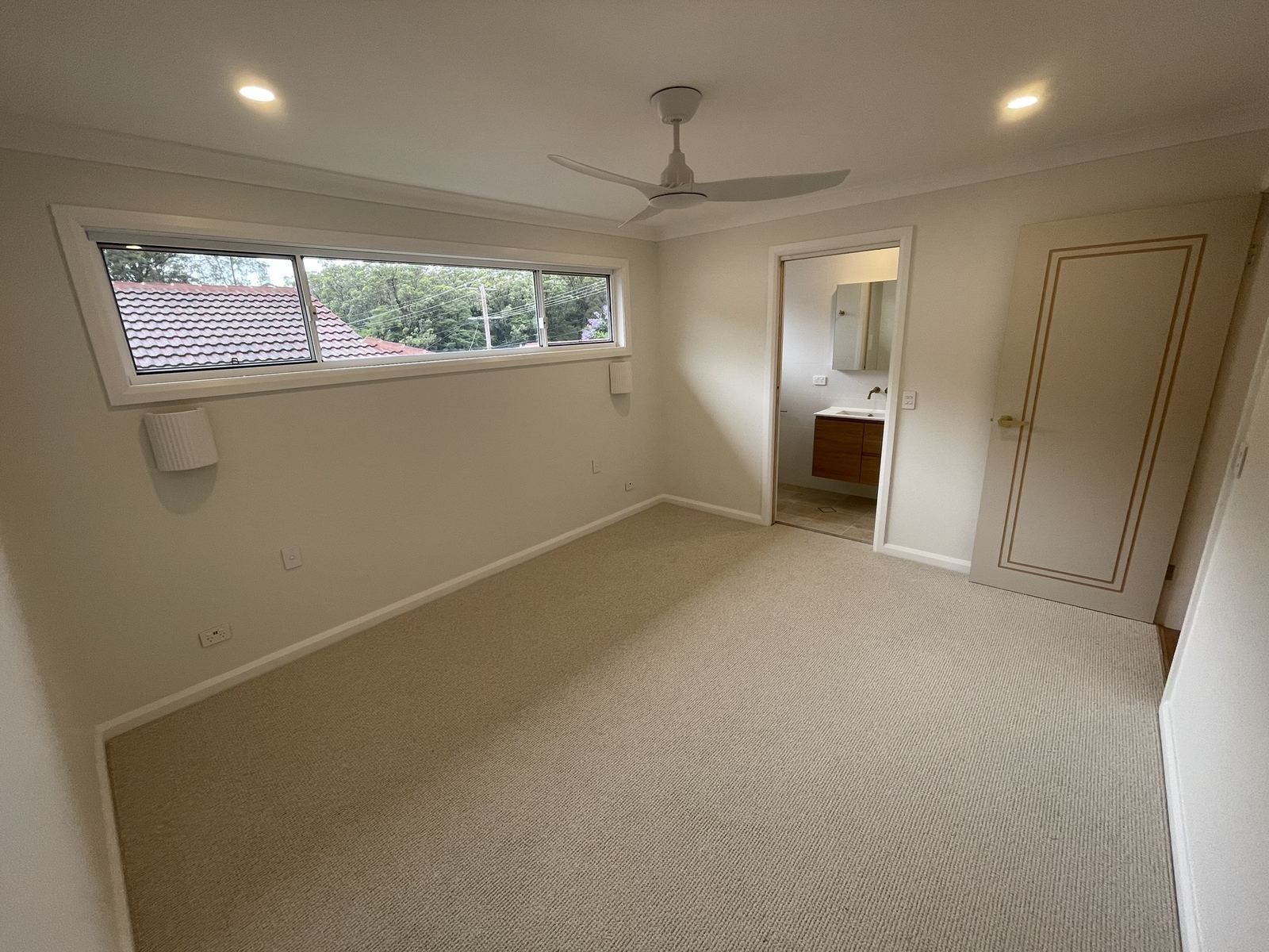 Empty beige bedroom with carpet, ceiling fan, long window, open doorway, and built-in closet.