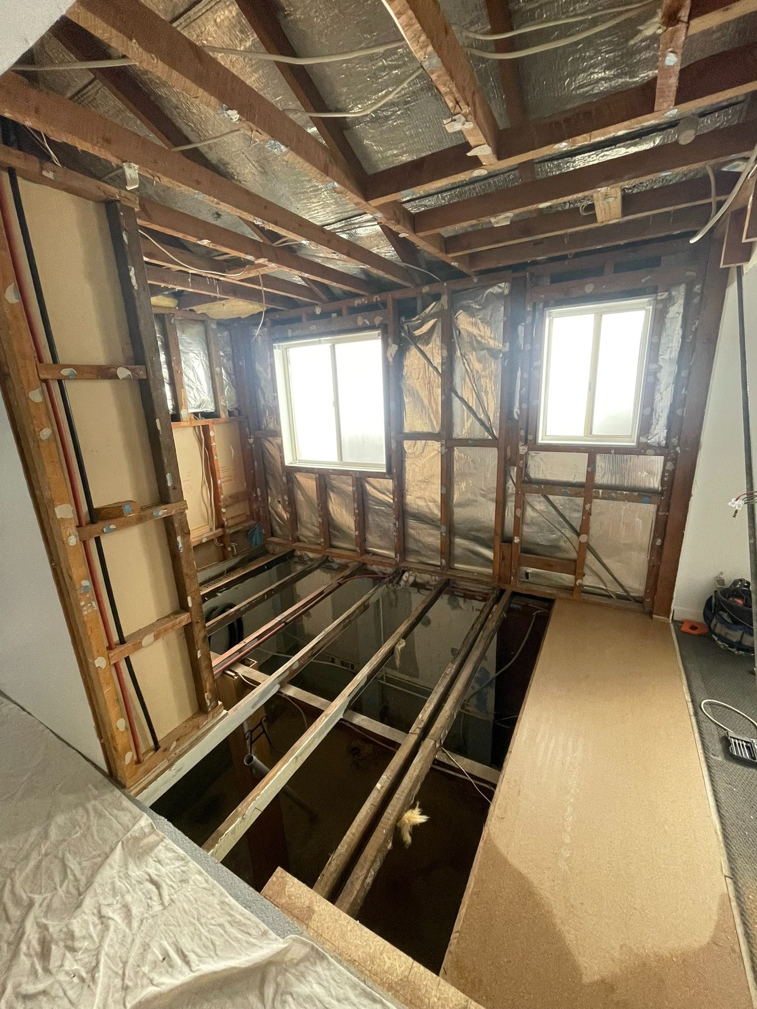 Exposed house framing and floor joists in a room under renovation, with daylight through two windows.