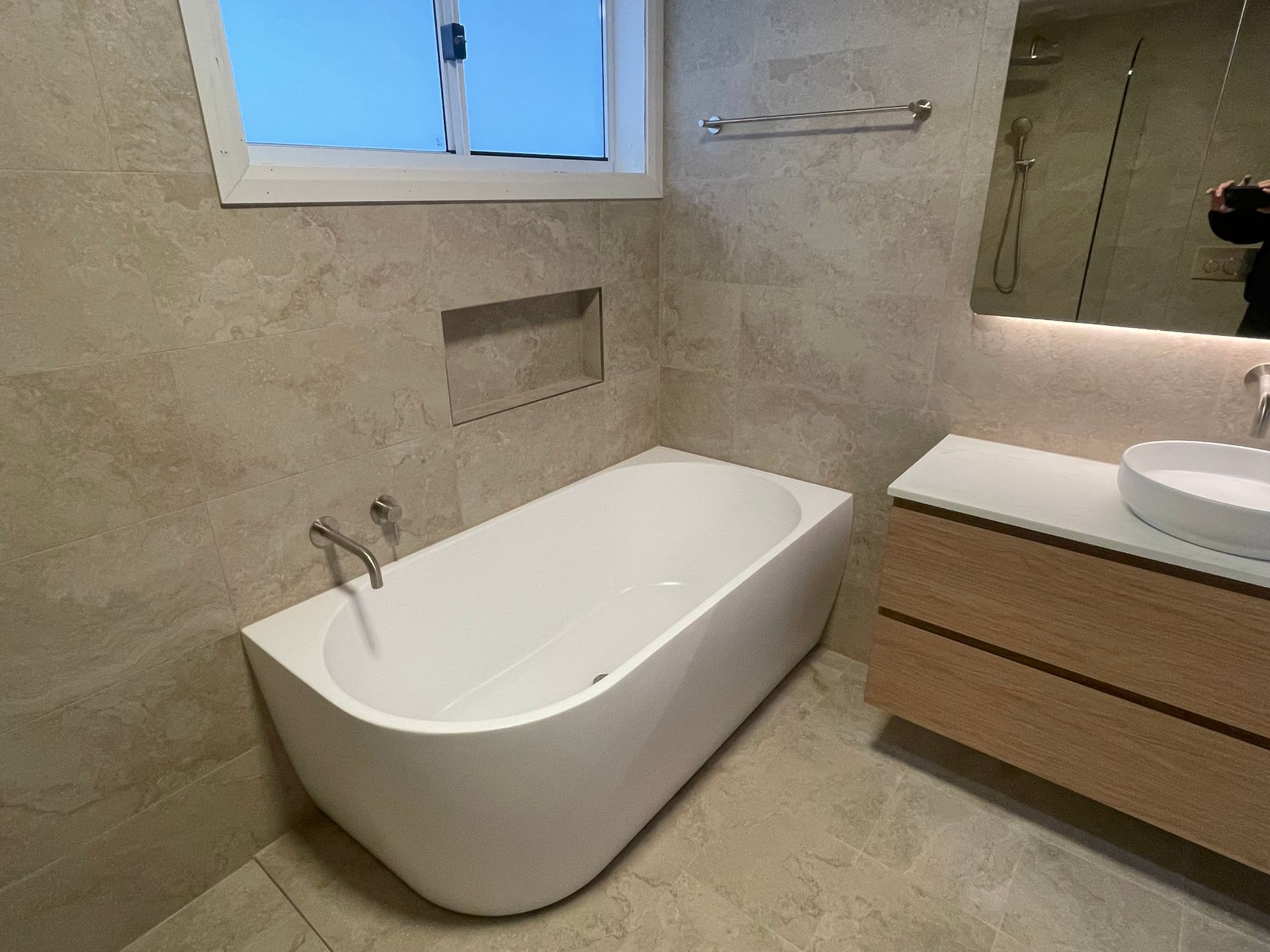 Modern bathroom with freestanding white tub, beige tile walls, and wooden vanity with sink and mirror