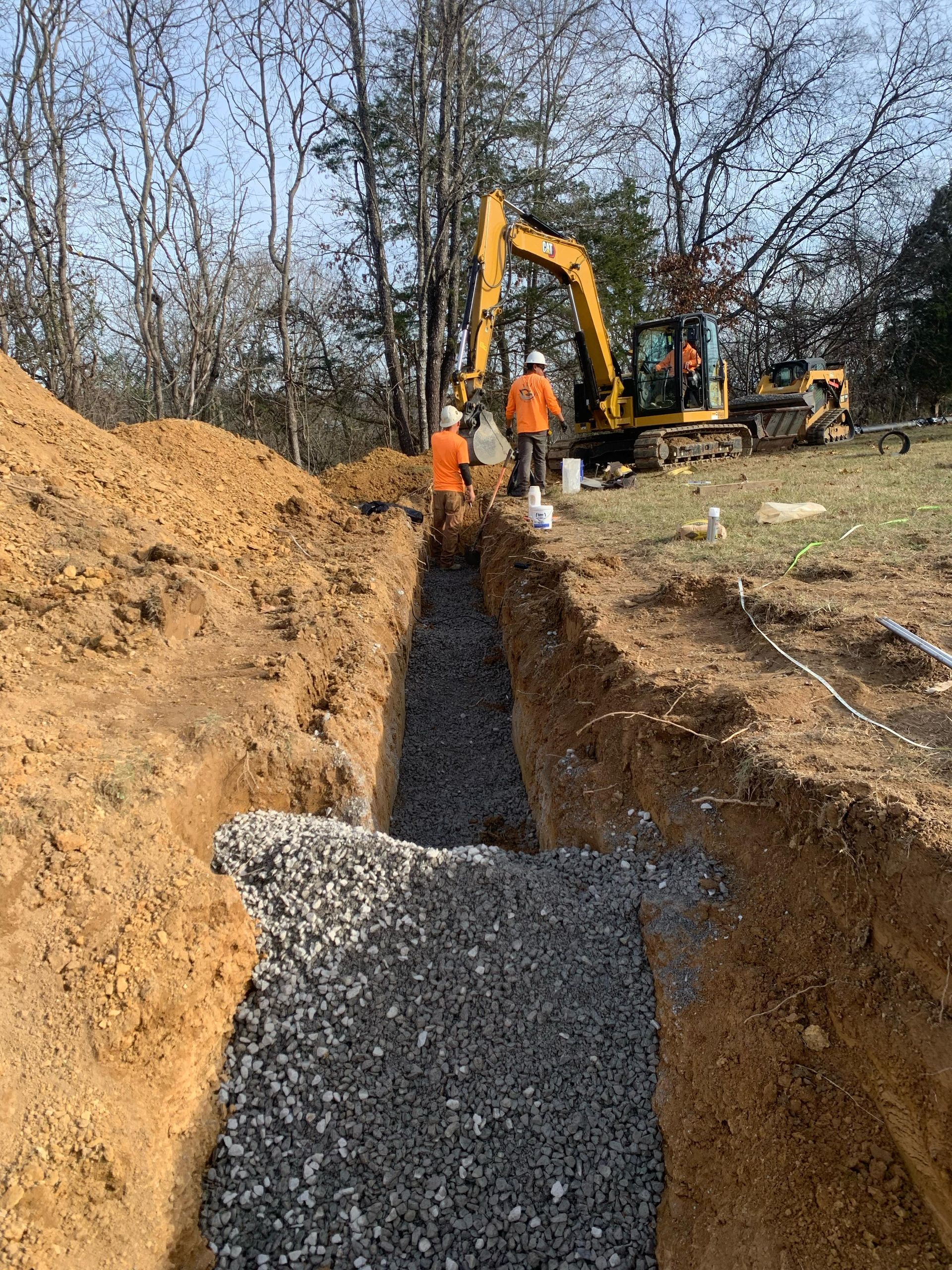 A yellow excavator is digging a trench in the dirt next to a pile of gravel.