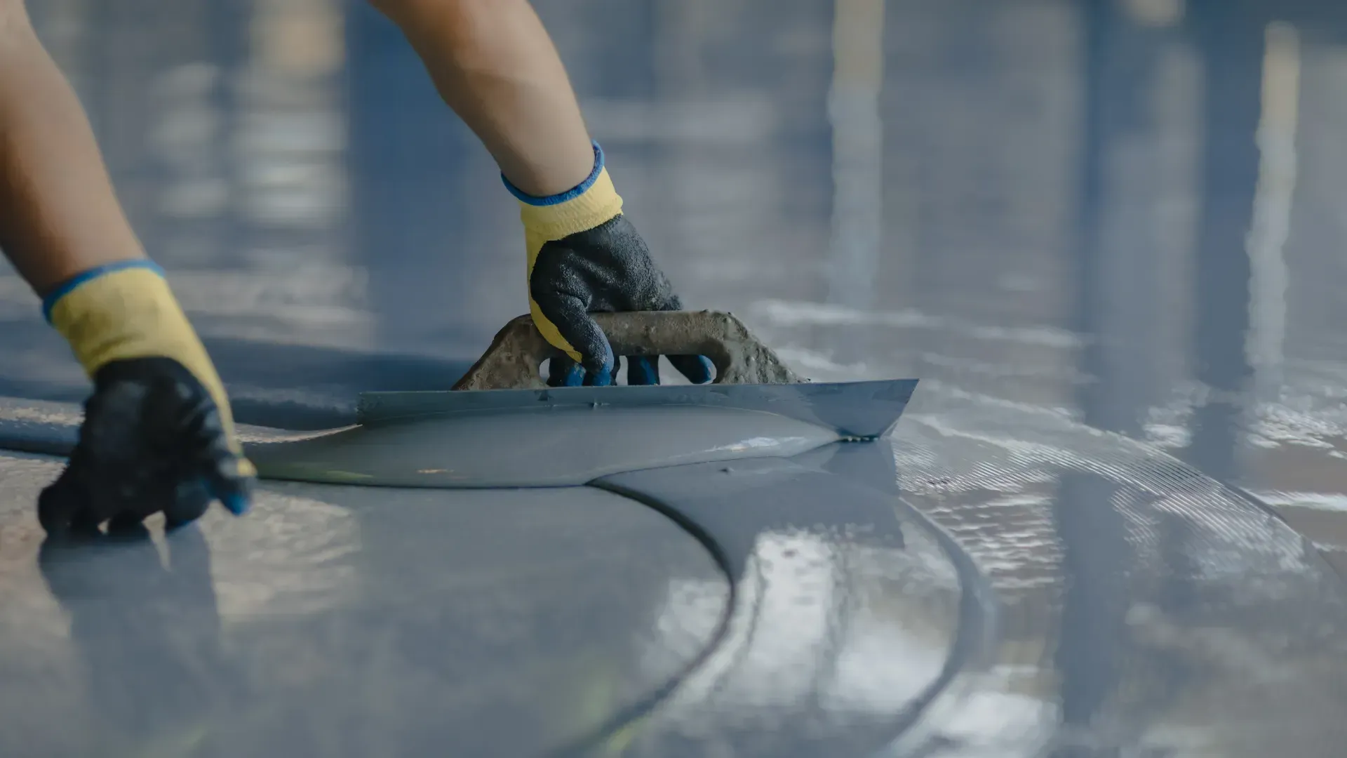 Hands wearing work gloves spread grey, liquid epoxy coating onto a floor using a trowel.