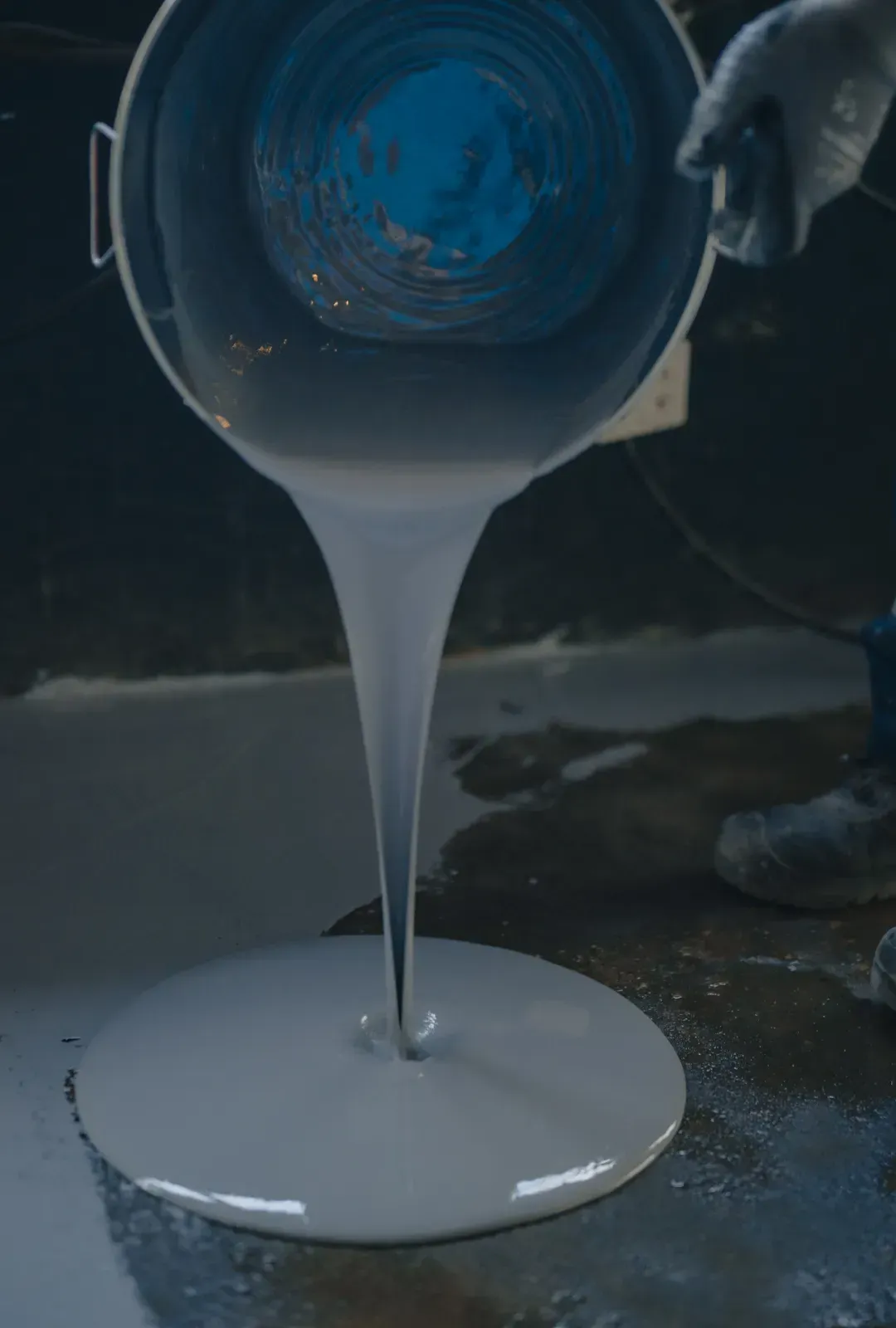 A gloved hand pours thick, creamy white liquid from a blue bucket onto a dark floor.