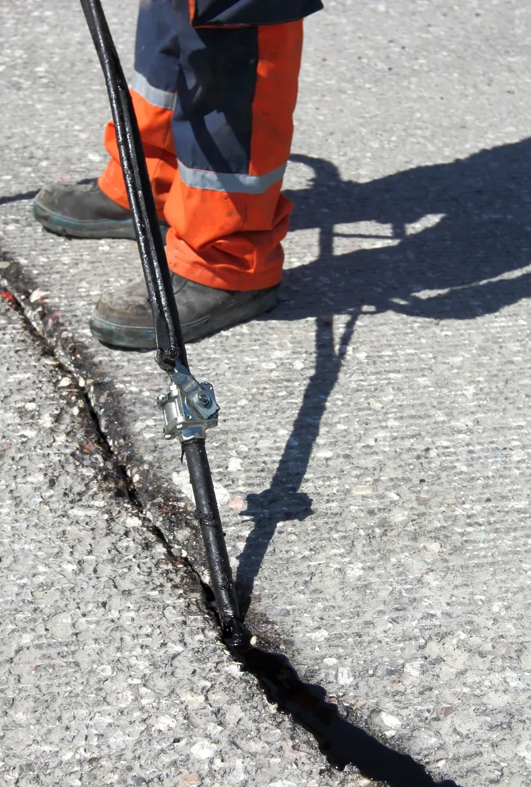 A worker in high-visibility orange pants fills a pavement crack with hot liquid sealant using a handheld applicator.
